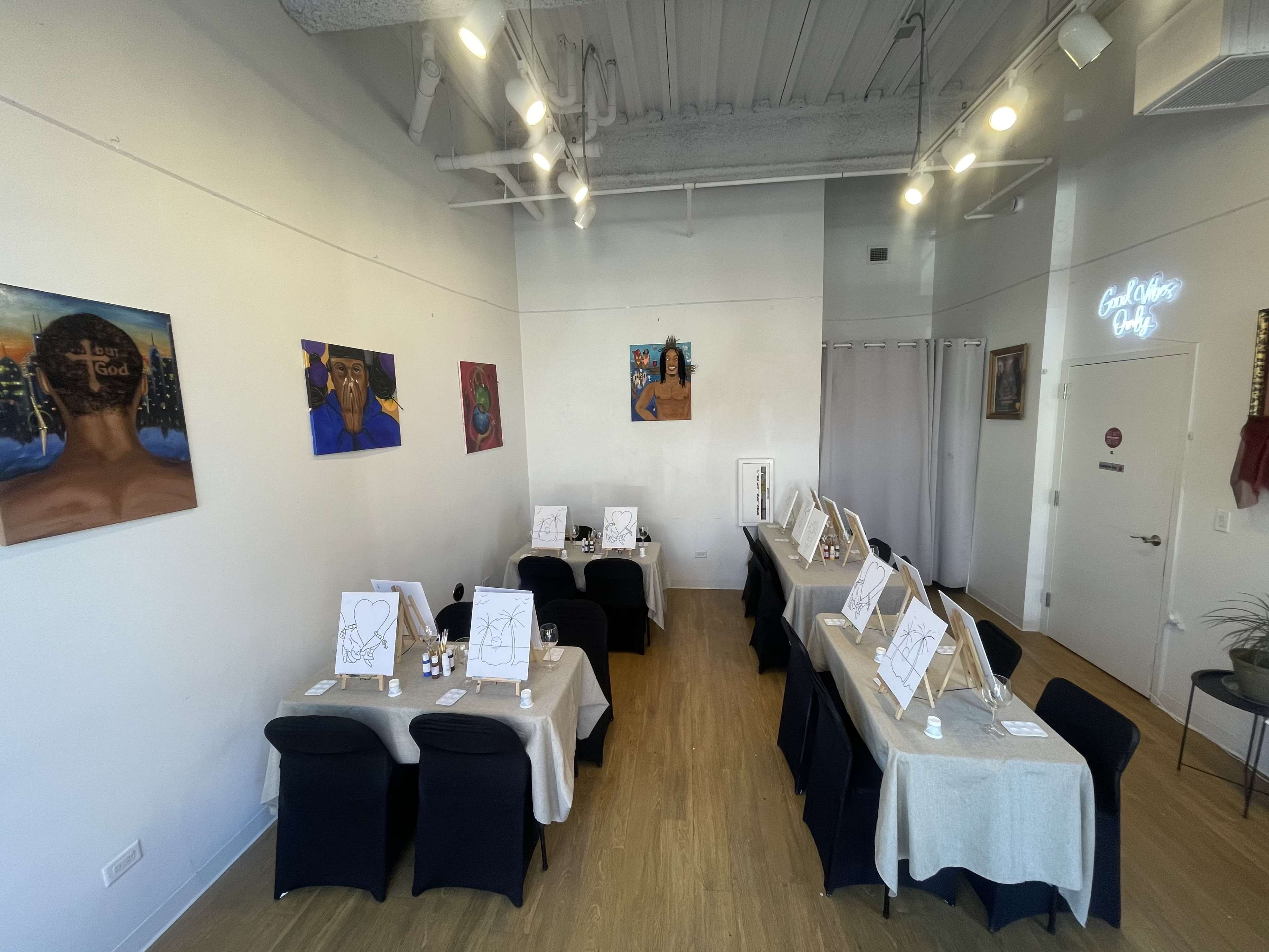 The image shows an art studio setup with multiple tables covered in beige tablecloths, each featuring a white drawing pad and art supplies, surrounded by colorful wall paintings.