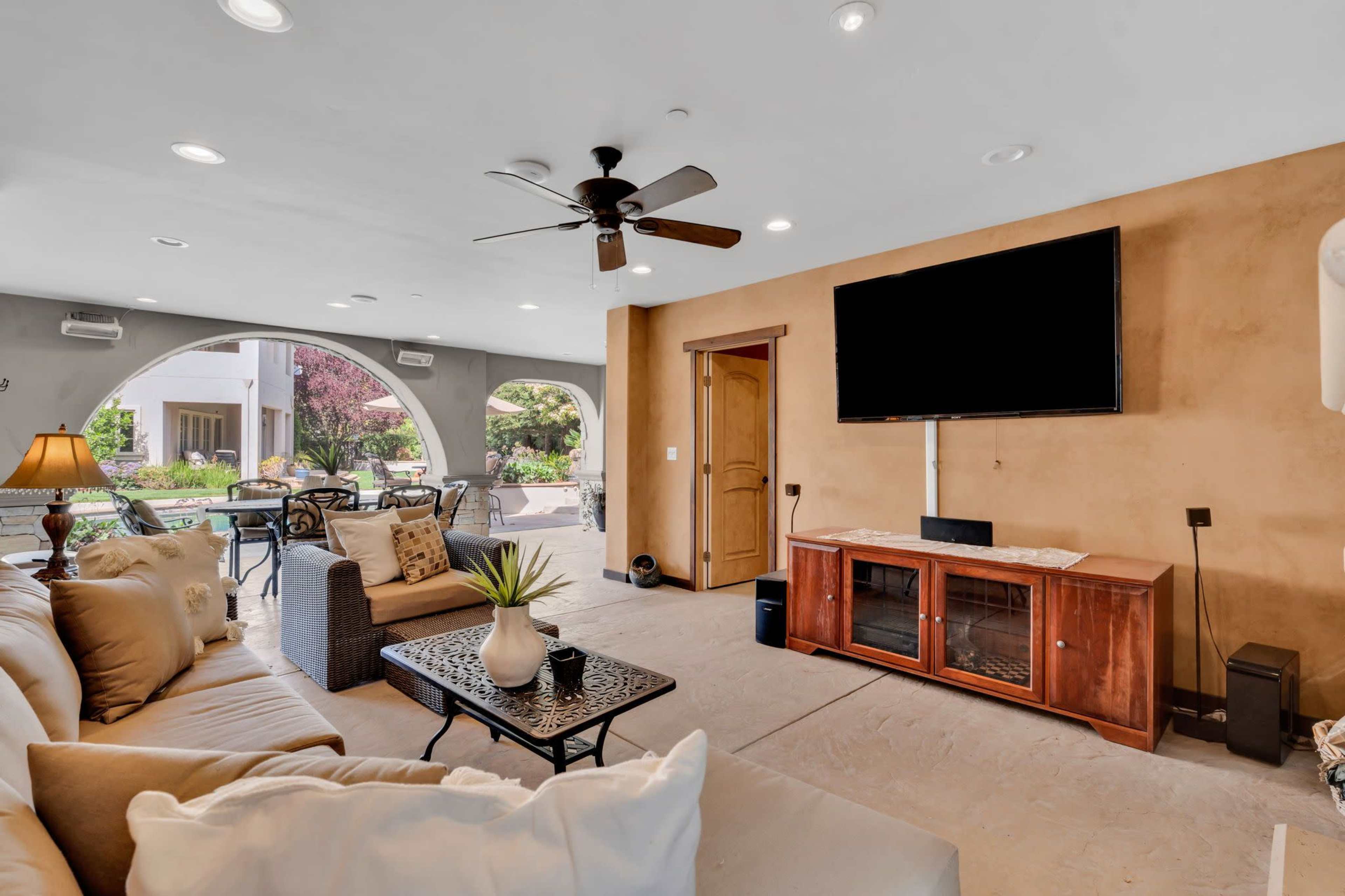 The image shows a spacious living room with a beige couch, a coffee table, a television mounted on the wall, and large arched openings leading to an outdoor seating area.