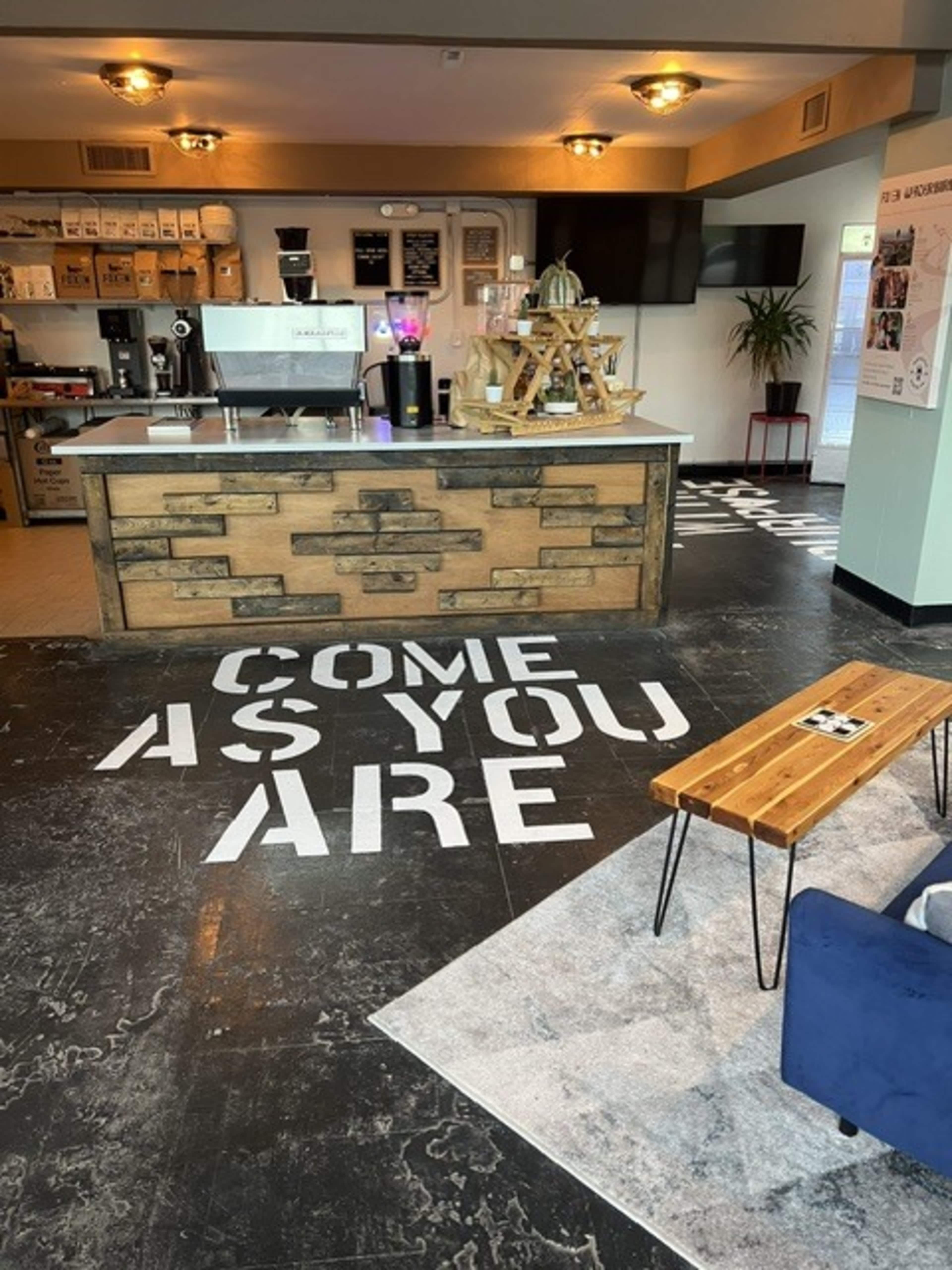 The image shows a modern café interior with a wooden counter, a coffee machine, and a floor featuring the phrase "COME AS YOU ARE."