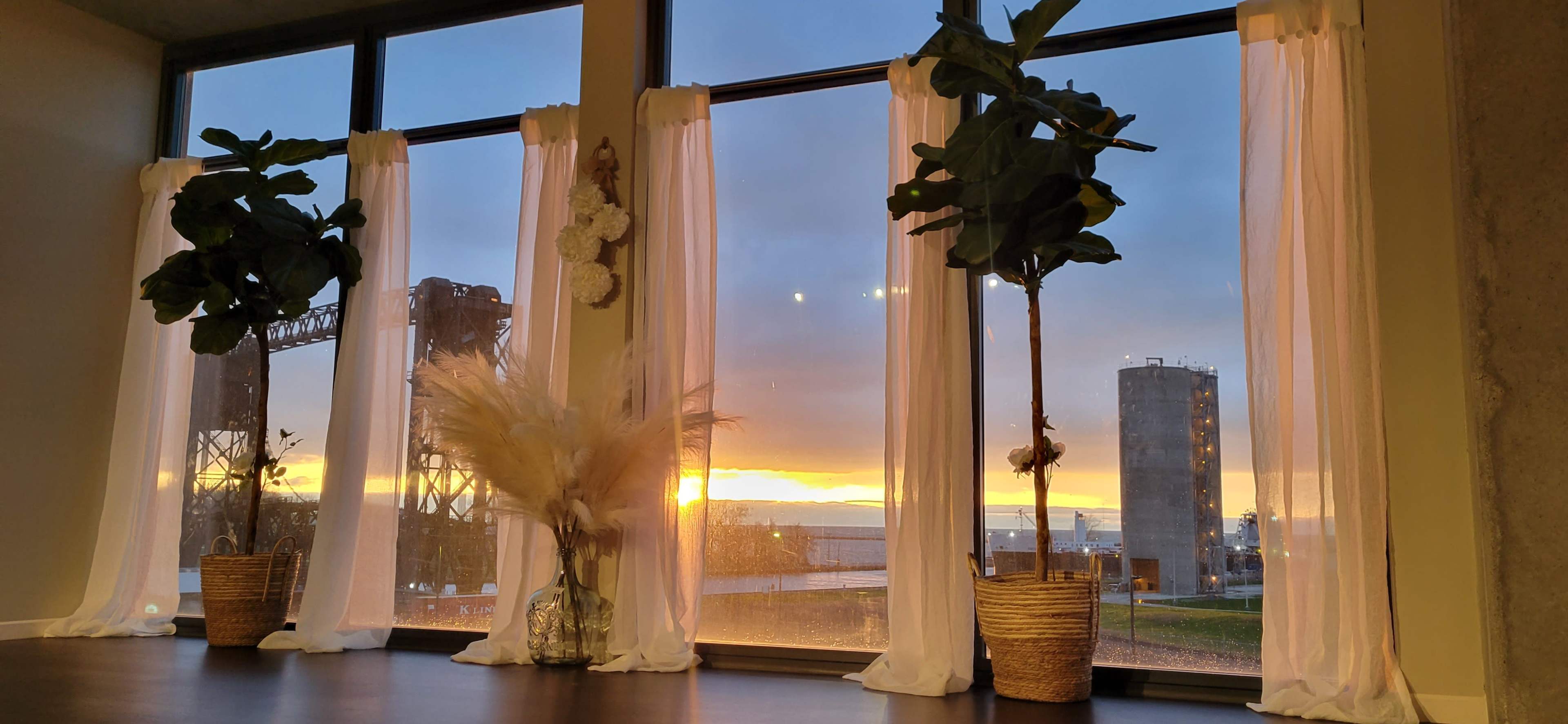 The scene shows a room with large windows revealing a sunset, framed by white curtains and featuring potted plants and decorative elements in front.