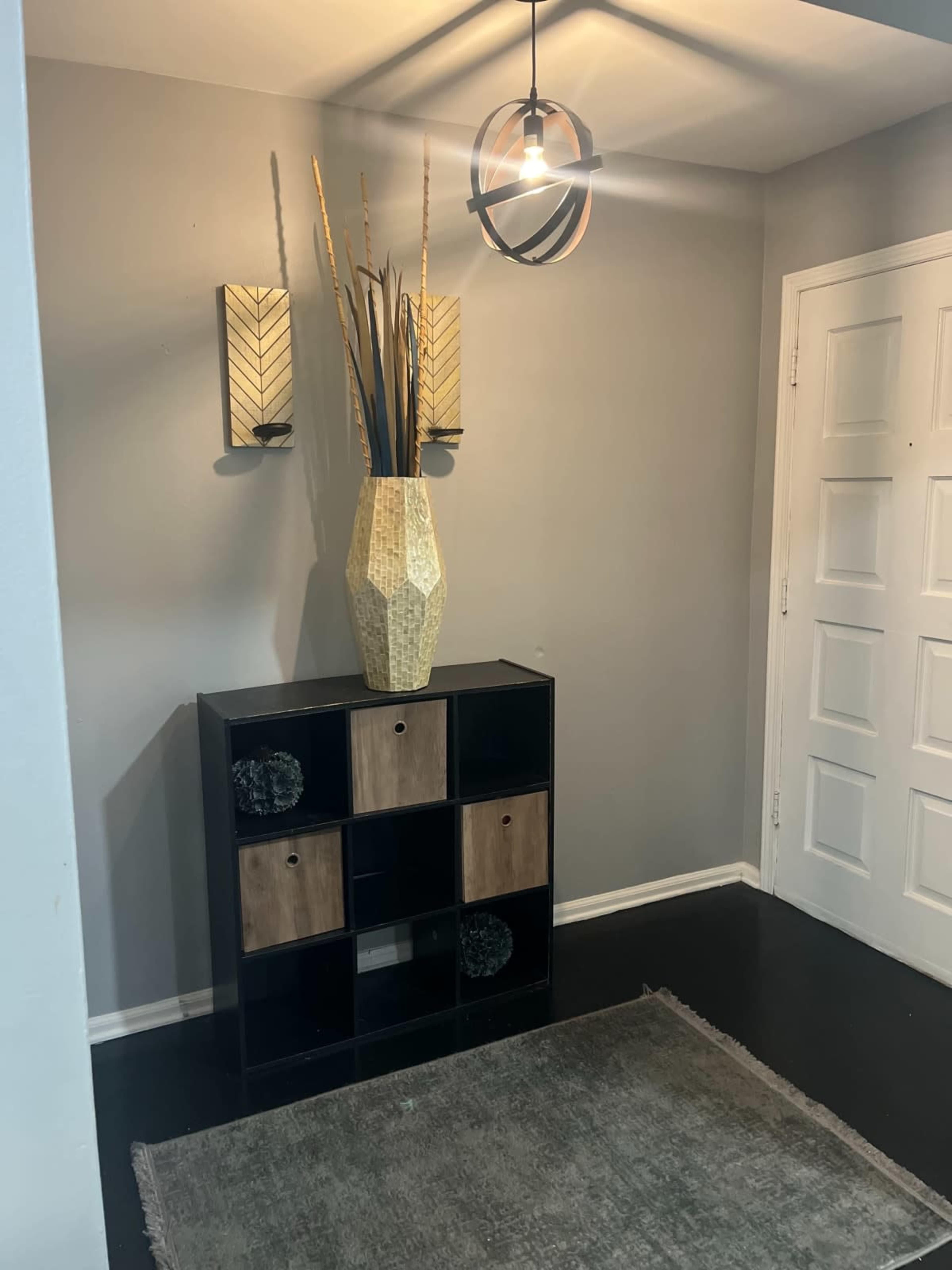 The image shows a small entryway with a black and wood cube storage unit, a tall decorative vase, wall sconces, and a door leading to another room.