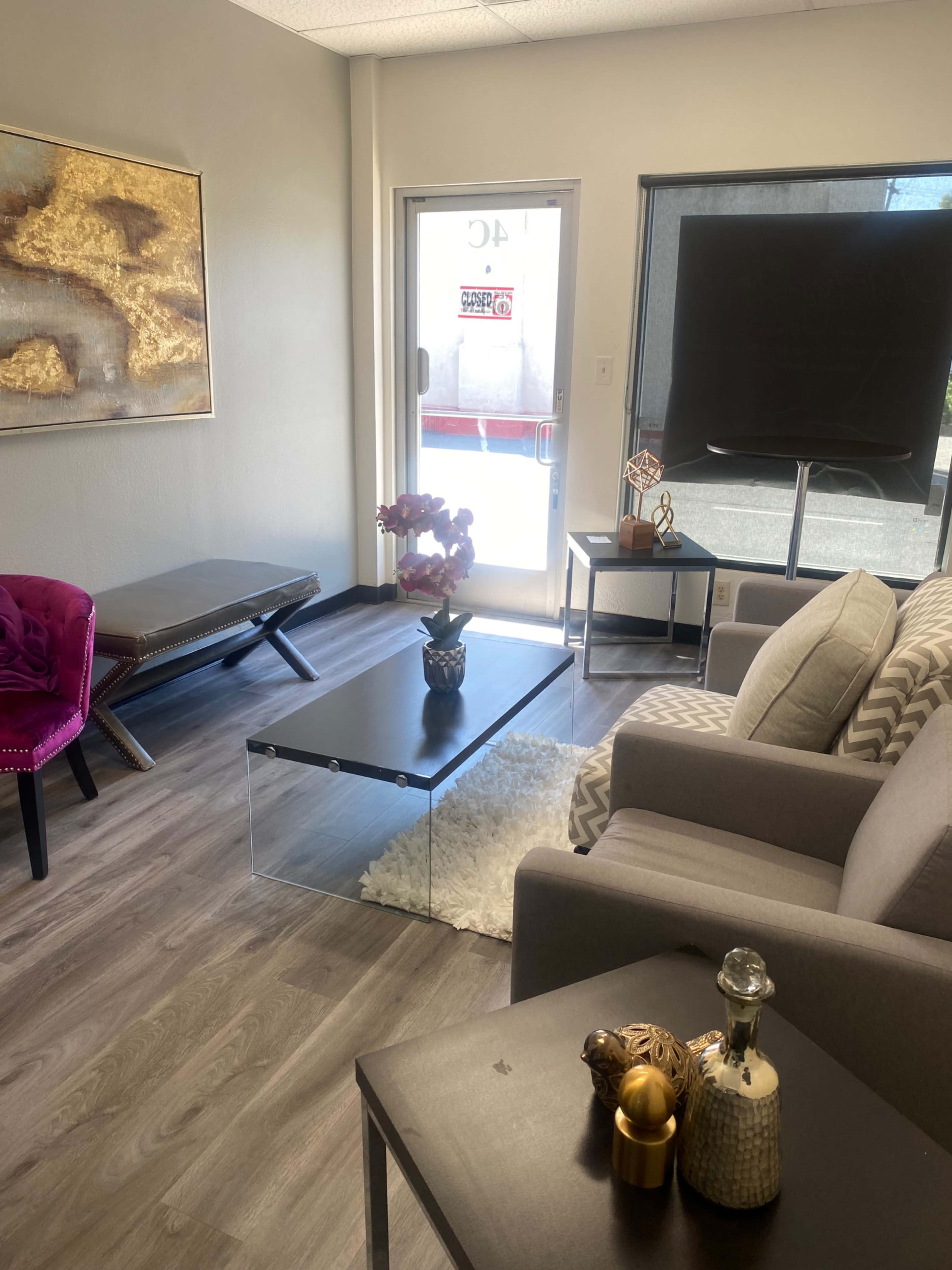 A modern waiting room with a glass coffee table, upholstered chairs, a large television, and a door leading outside.