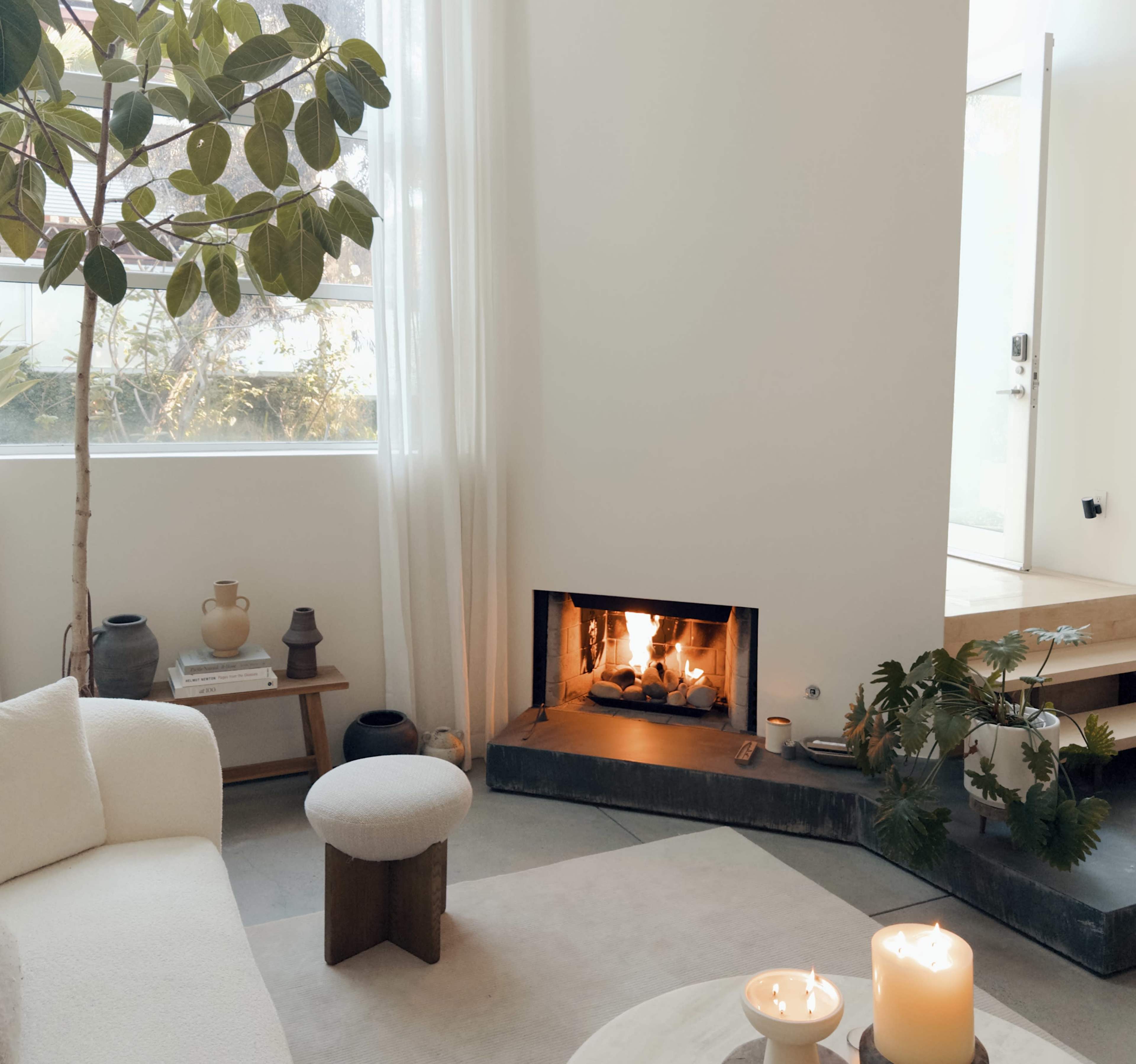 A cozy living room features a modern fireplace, a light-colored sofa, a small wooden stool, and indoor plants by large windows.