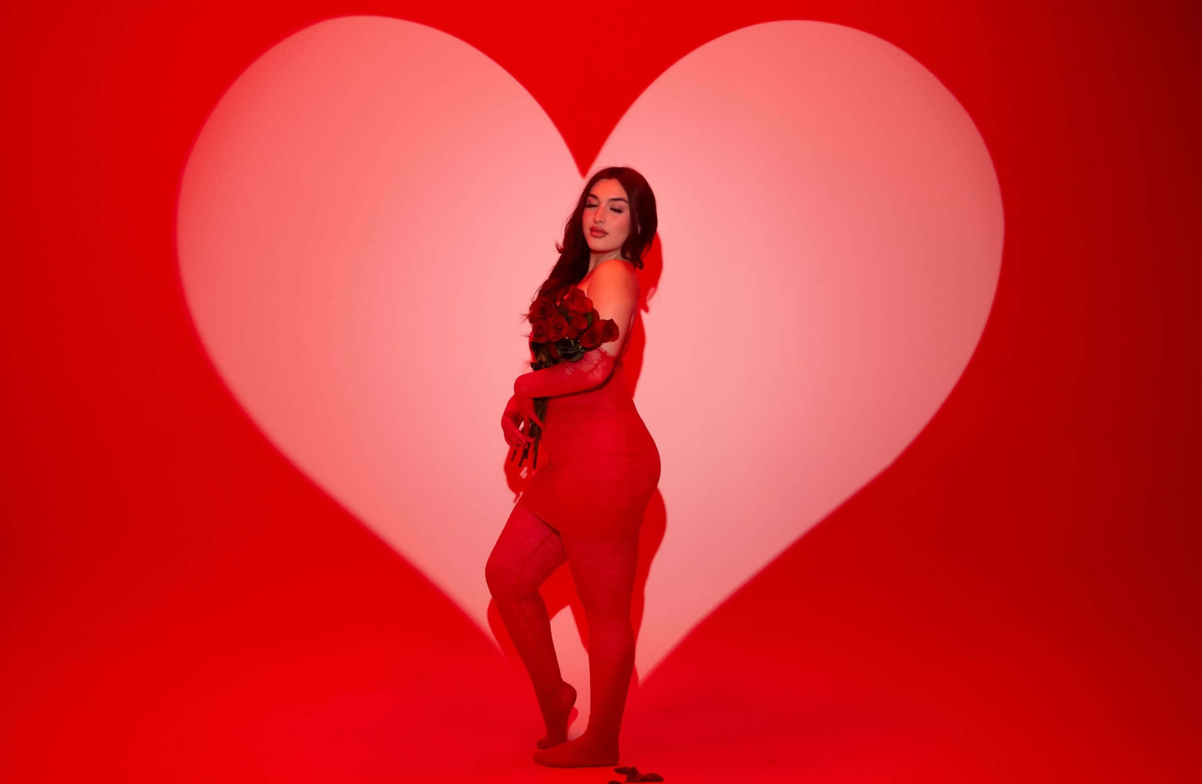 A woman in a red outfit holds a bouquet of roses in front of a large heart-shaped backdrop.