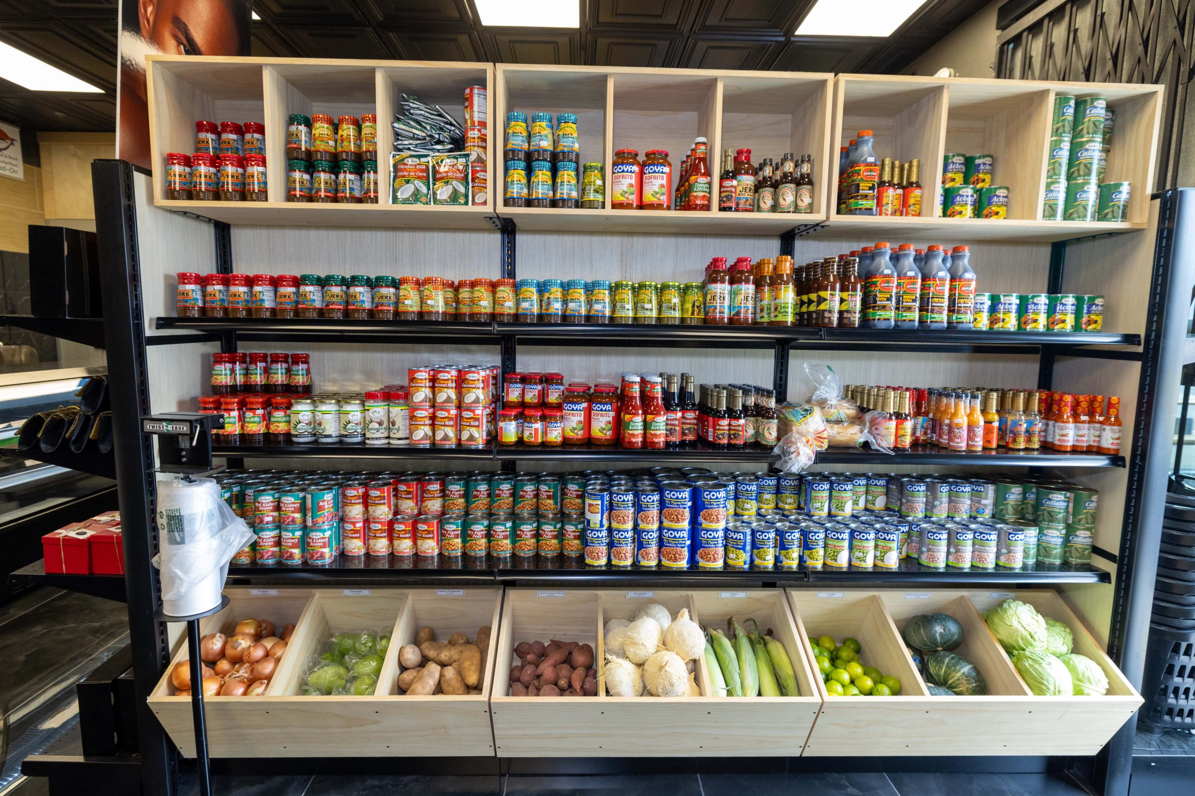 A grocery display features shelves stacked with various jars and cans of sauces, spices, and canned goods, alongside a lower section containing fresh vegetables and eggs.