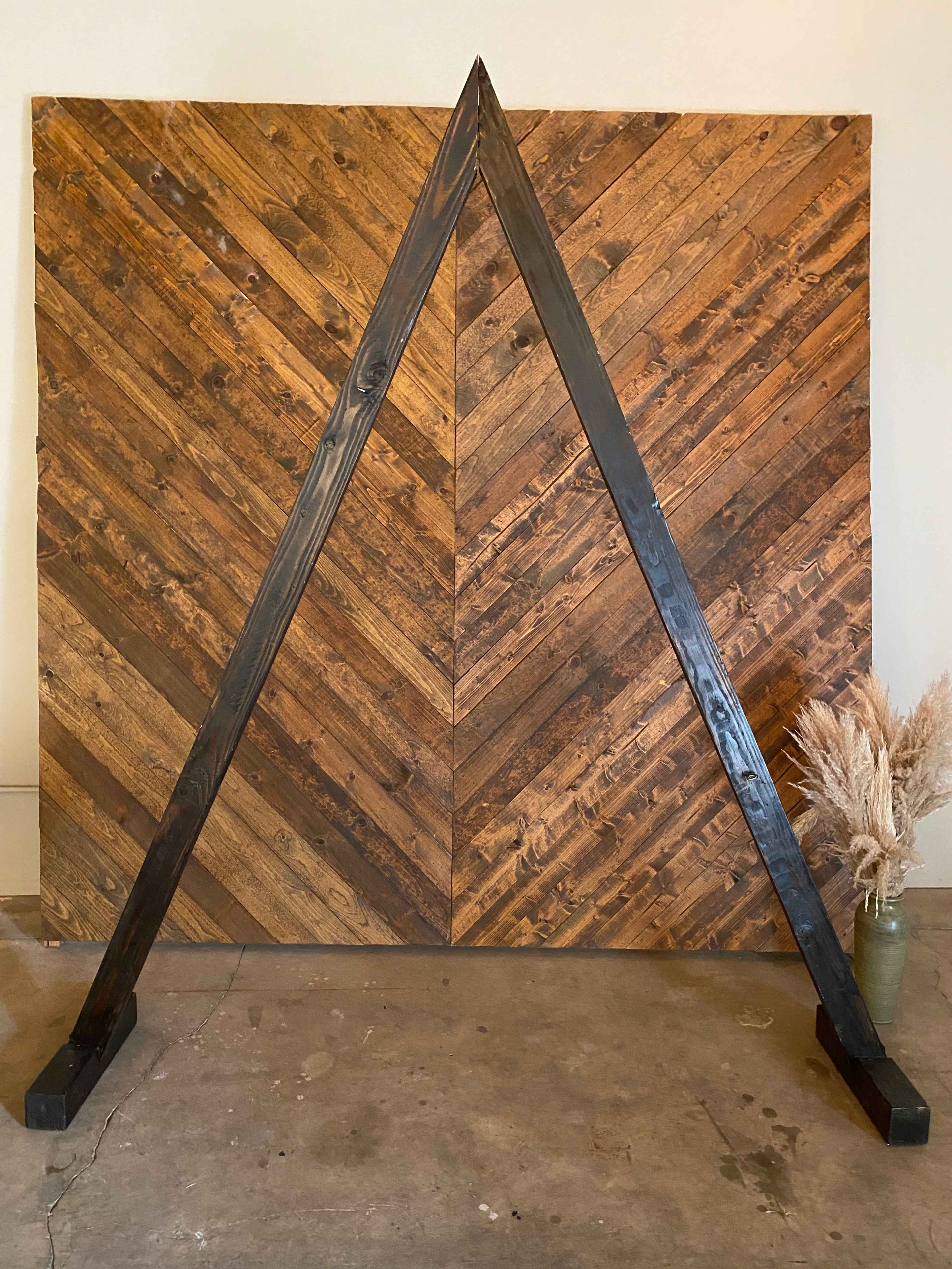 A large wooden frame stands in front of a herringbone-patterned wood backdrop, with dried plants in a vase positioned to the side.