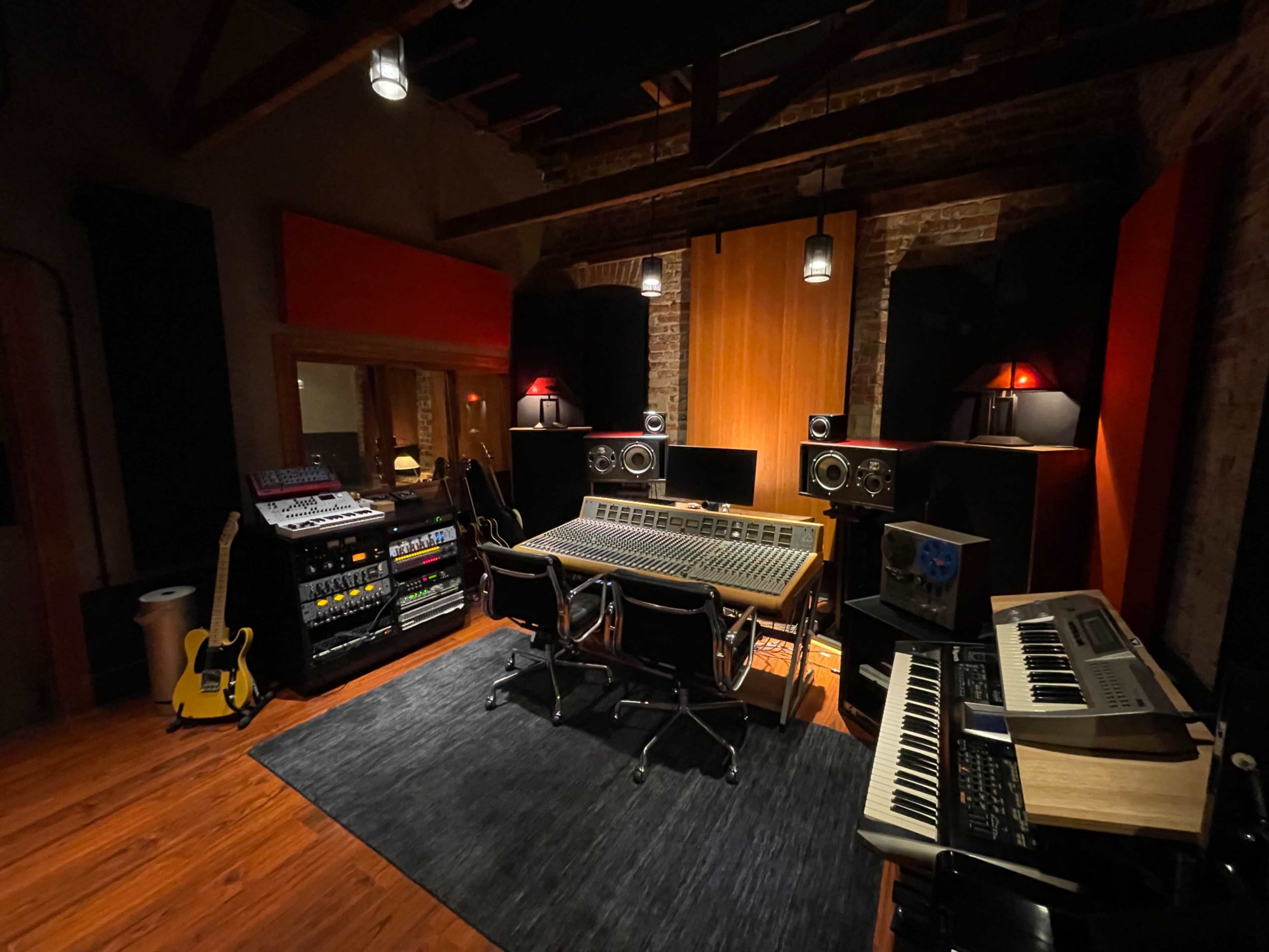A recording studio with a large mixing console, various audio equipment, guitars, and keyboards arranged in a well-lit space featuring exposed brick walls and wooden beams.