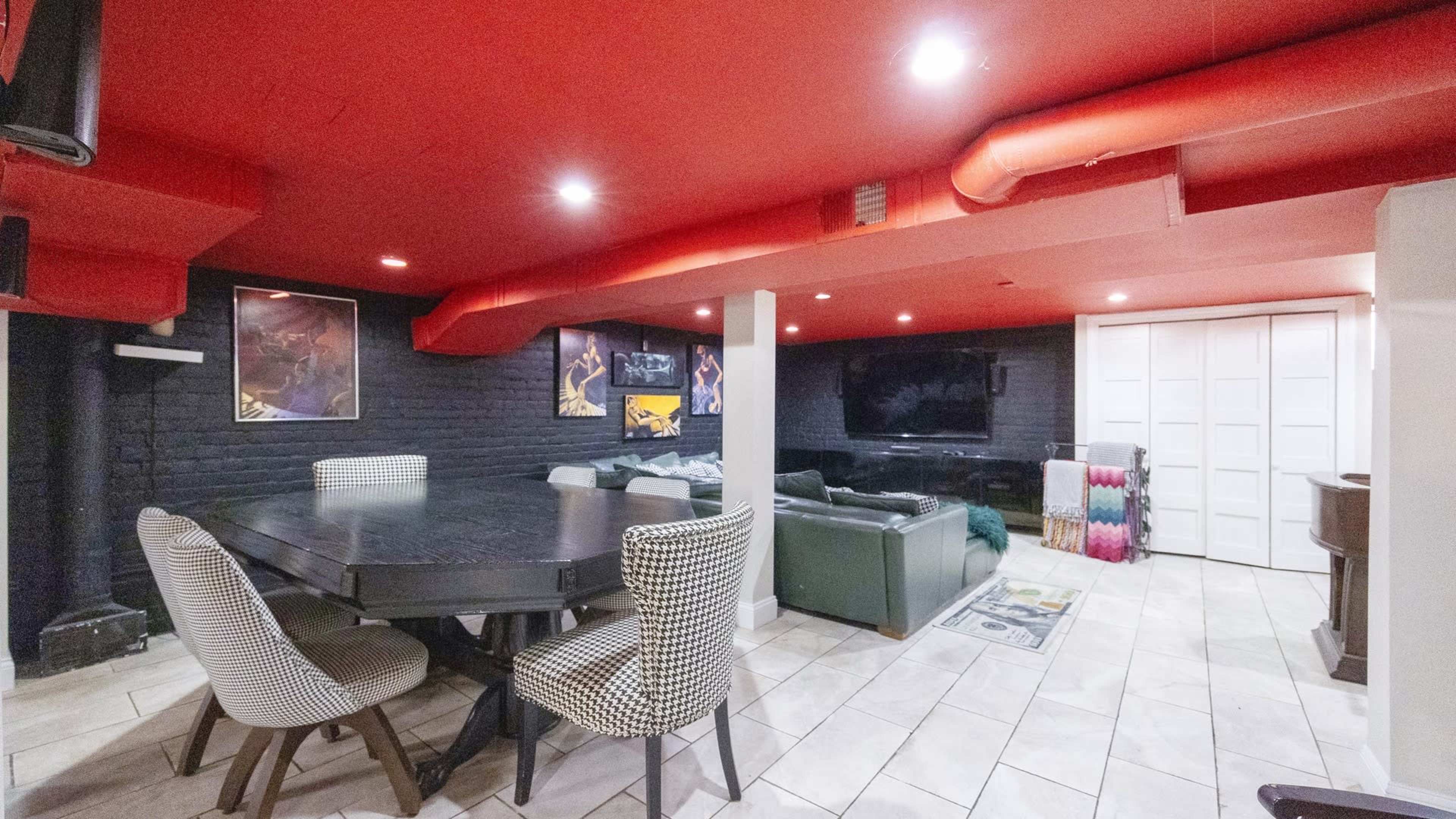 A modern basement with red ceilings, a large dining table with chairs, and a seating area featuring a green sofa and a television.