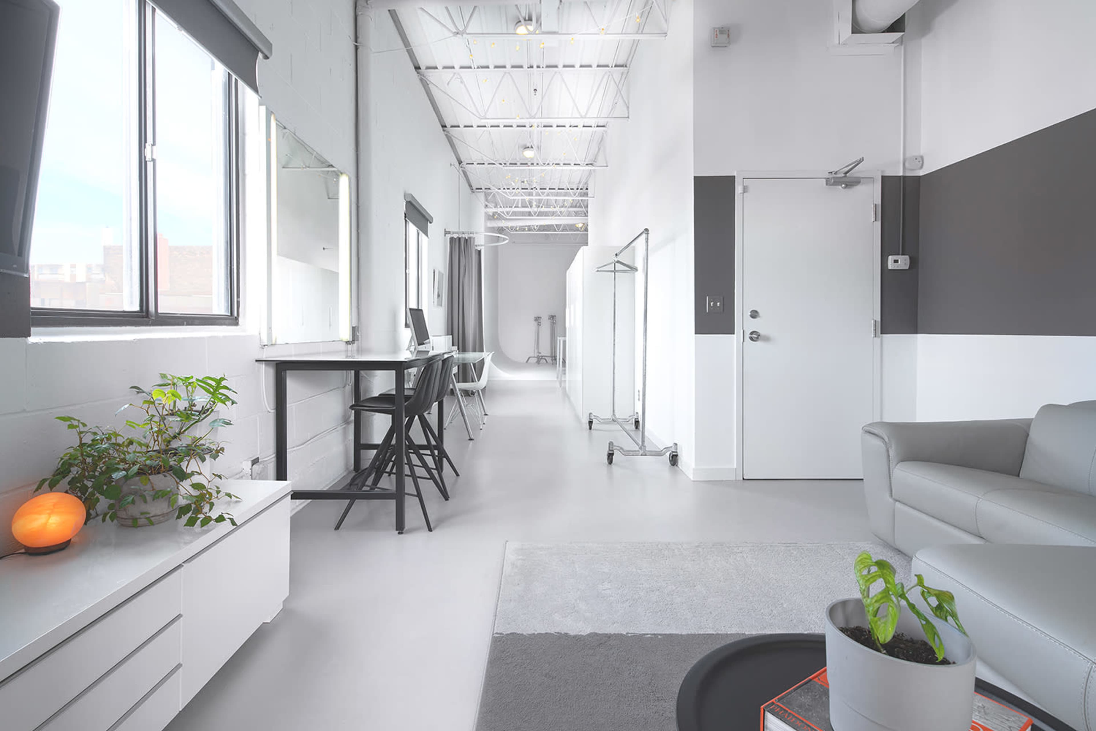 The image shows a bright, modern interior featuring a long hallway with large windows, a gray couch, a small plant, and a minimalist workspace along one side.