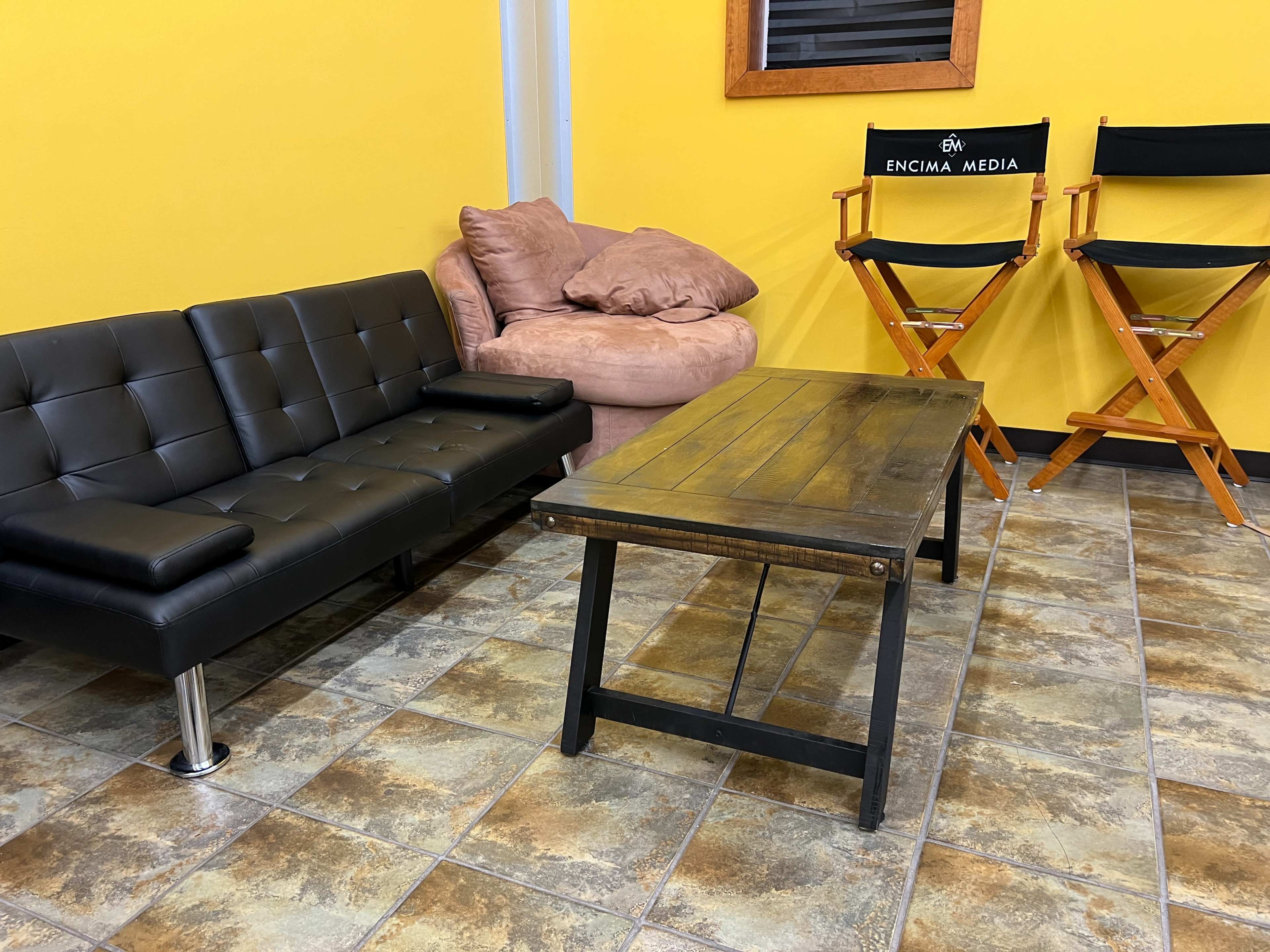 A room with a black leather couch, a pink armchair, a wooden table, and two director's chairs against a yellow wall.