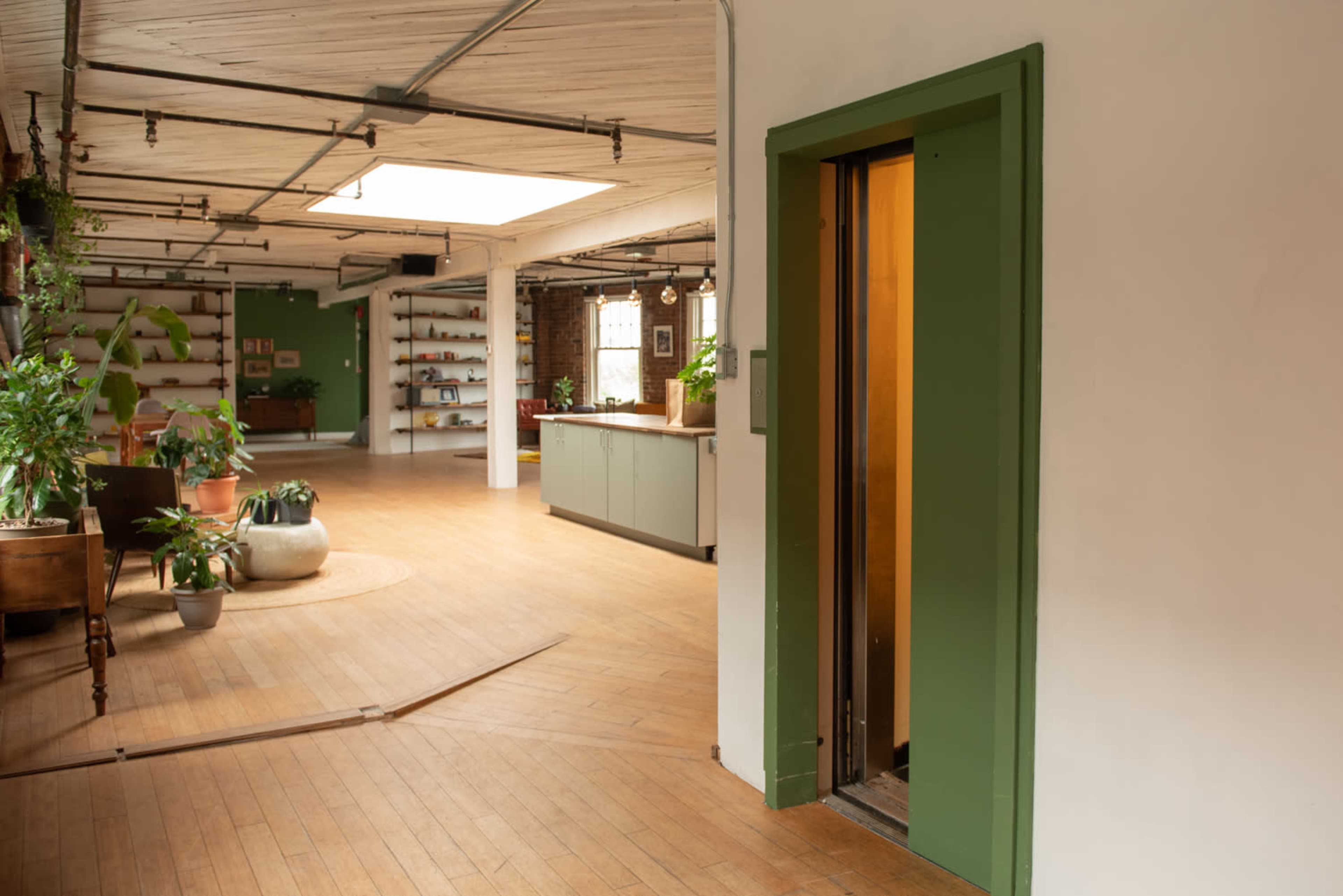 A spacious interior with wooden flooring, a green elevator door, and various plants throughout the room.
