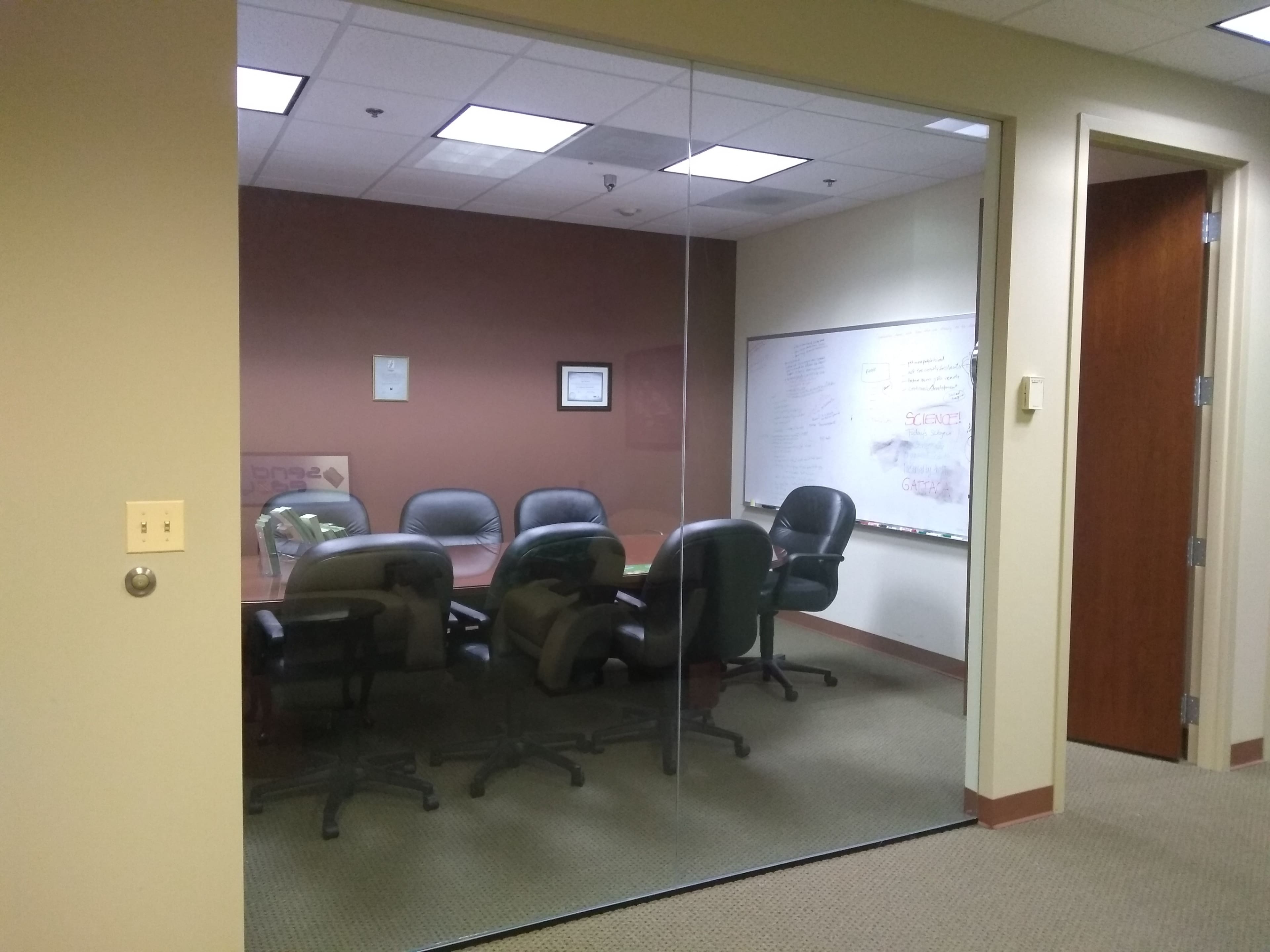 A conference room with a glass wall, featuring a large table surrounded by six black chairs and a whiteboard covered in writing.