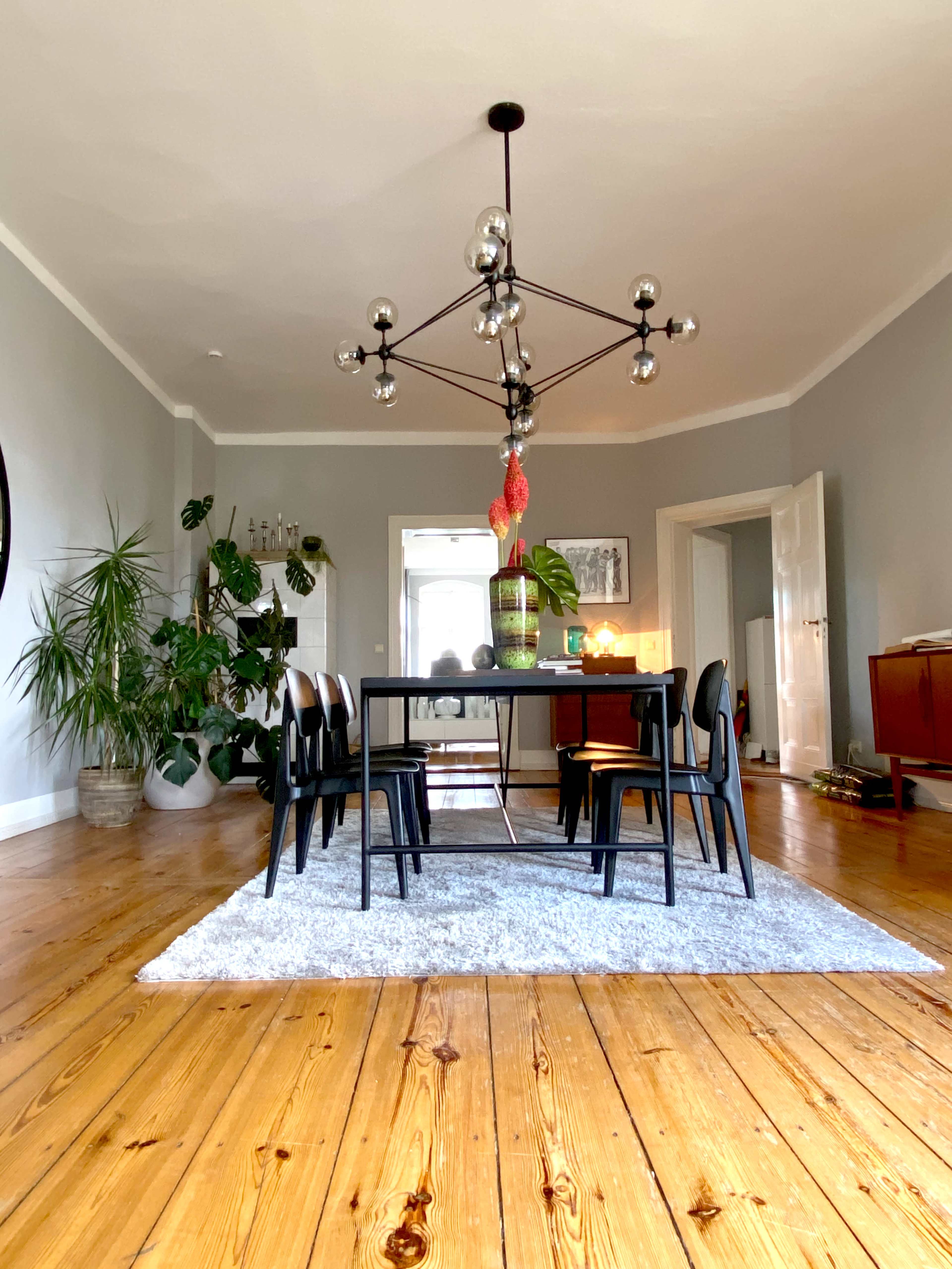 A dining area features a rectangular table surrounded by six black chairs on a light gray rug, with a large chandelier overhead and various plants in the background.