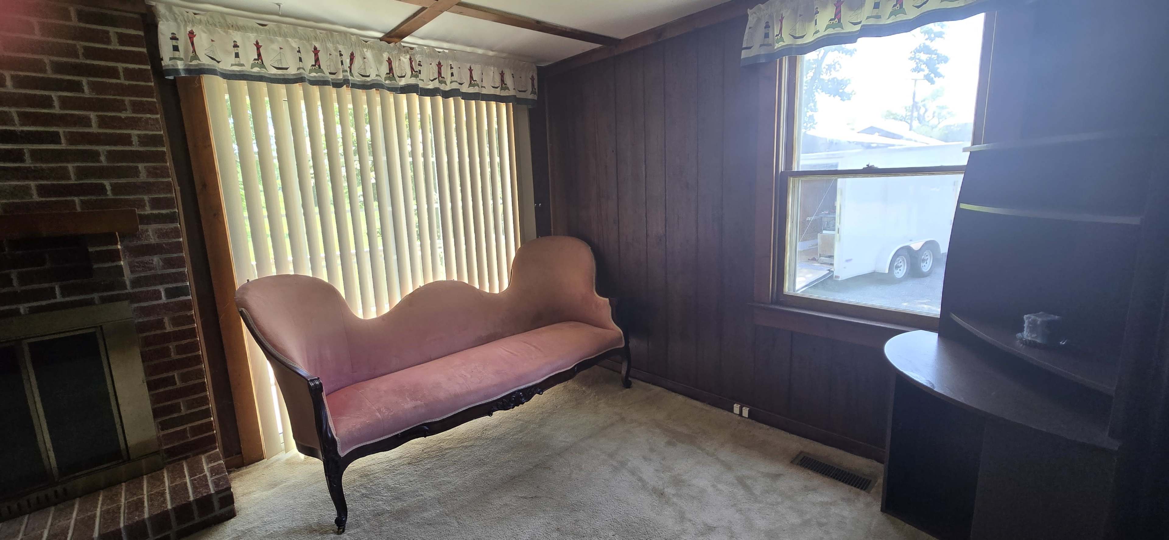 The image shows a modest room with a pink upholstered sofa, a brick fireplace, and a window covered by vertical blinds.