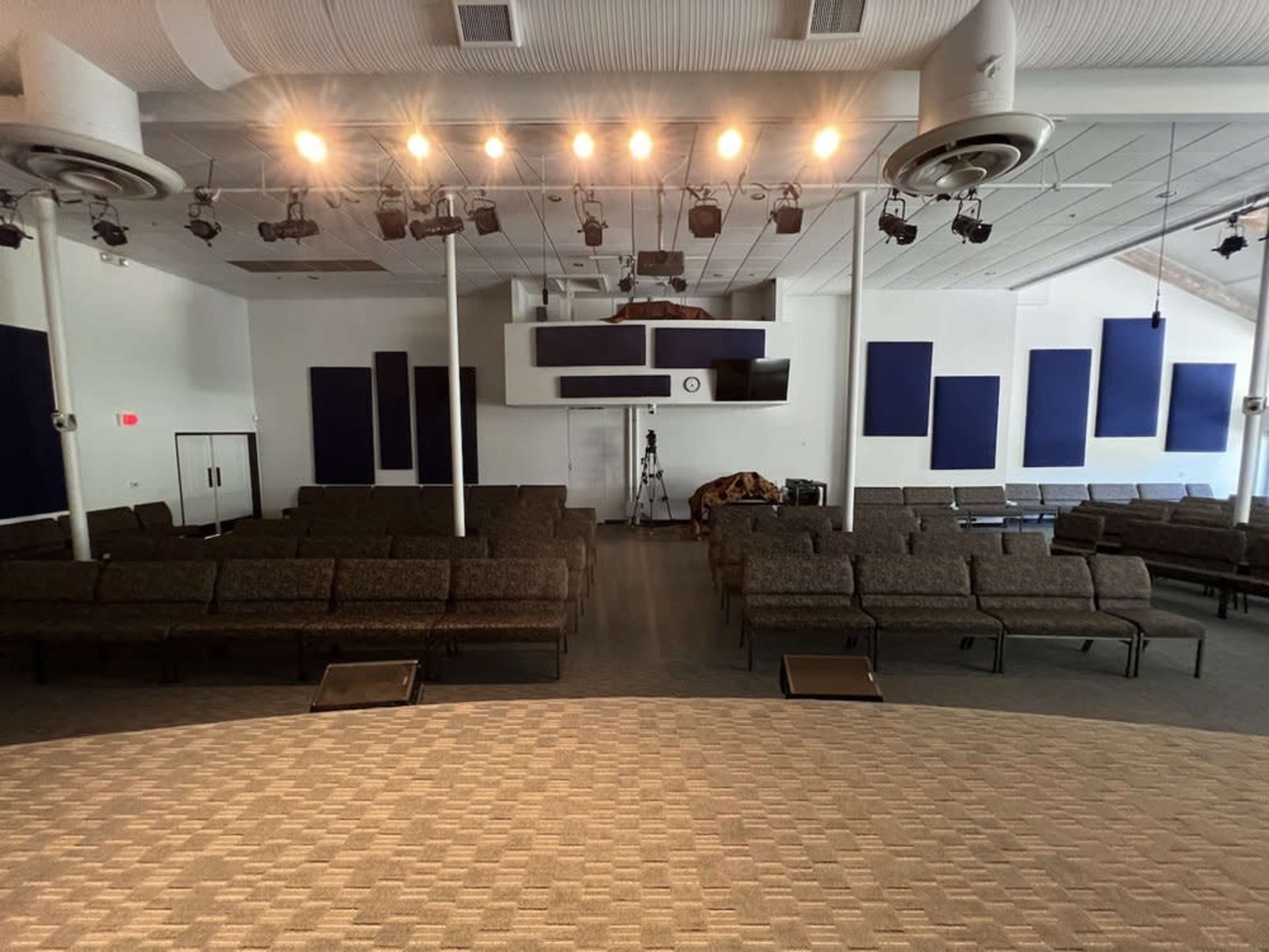 An empty auditorium with rows of chairs facing a stage, featuring overhead lights and sound equipment.