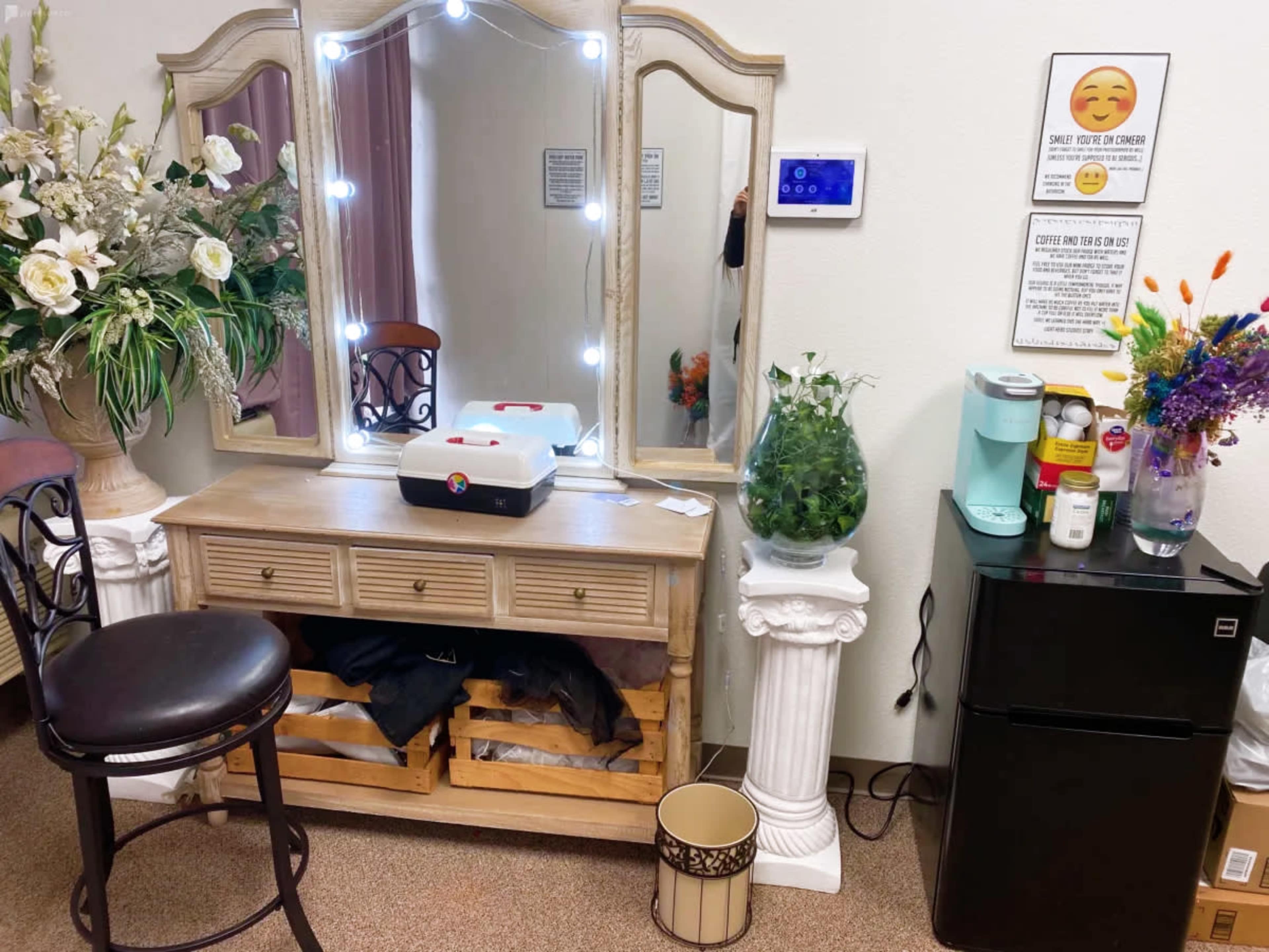 The image shows a mirrored vanity with a lighted mirror, a wooden cabinet, a small black stool, and decorative plants alongside a coffee maker and a mini fridge in a room.