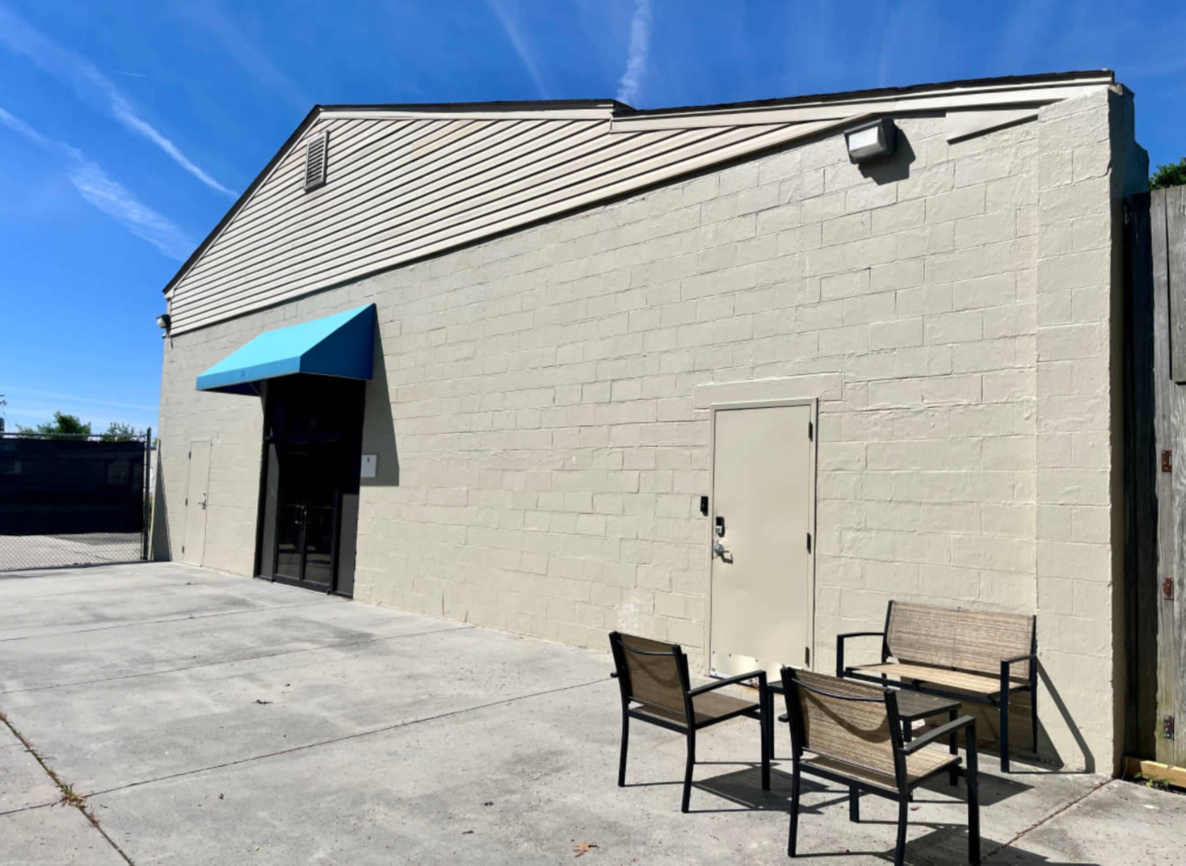 The image shows a light-colored building with an awning, a small door, and two chairs positioned on a concrete area outside.
