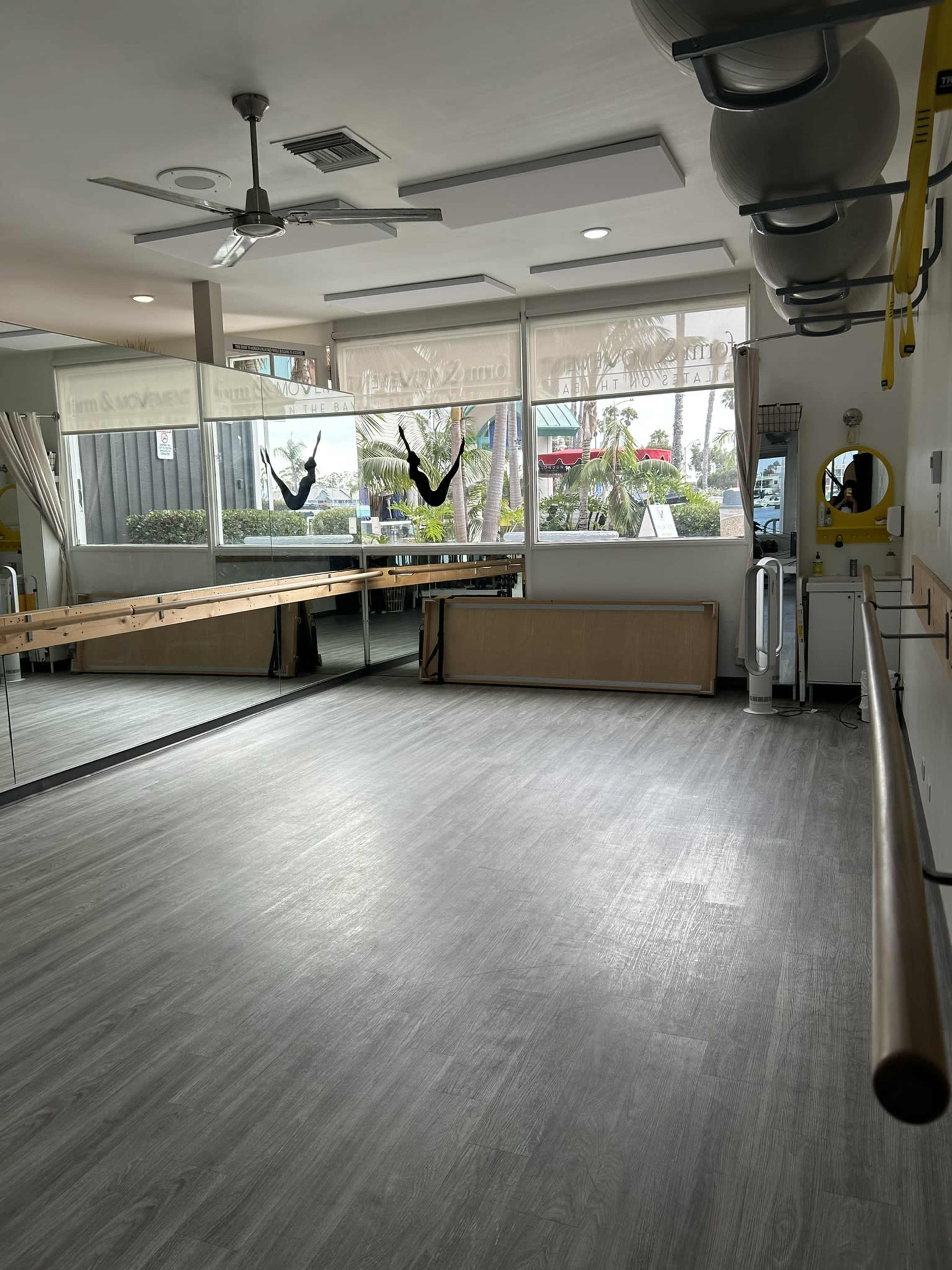 The image shows a light-filled room with large windows, a ballet barre along one wall, and mirrors reflecting the space, set up for fitness or dance activities.