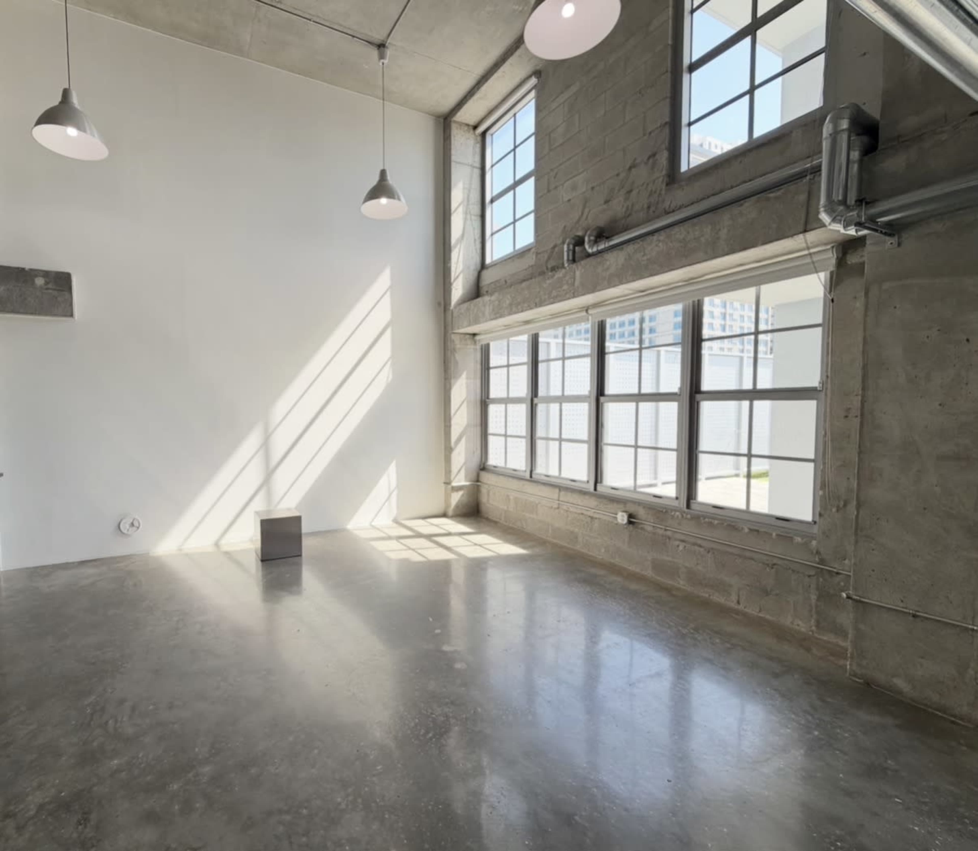 The image shows a spacious, empty room with large windows allowing sunlight to stream in, illuminating the polished concrete floor.