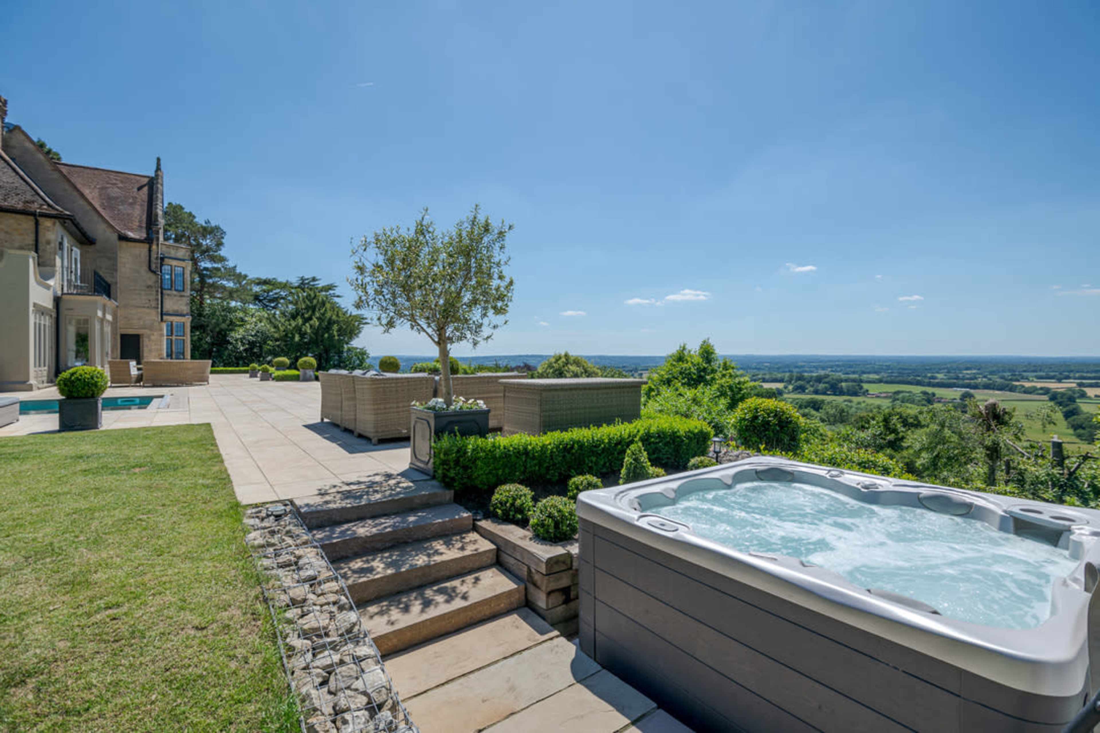 A backyard scene features a hot tub on a stone patio, surrounded by neatly trimmed hedges and steps leading to a lush green landscape with distant hills under a clear blue sky.