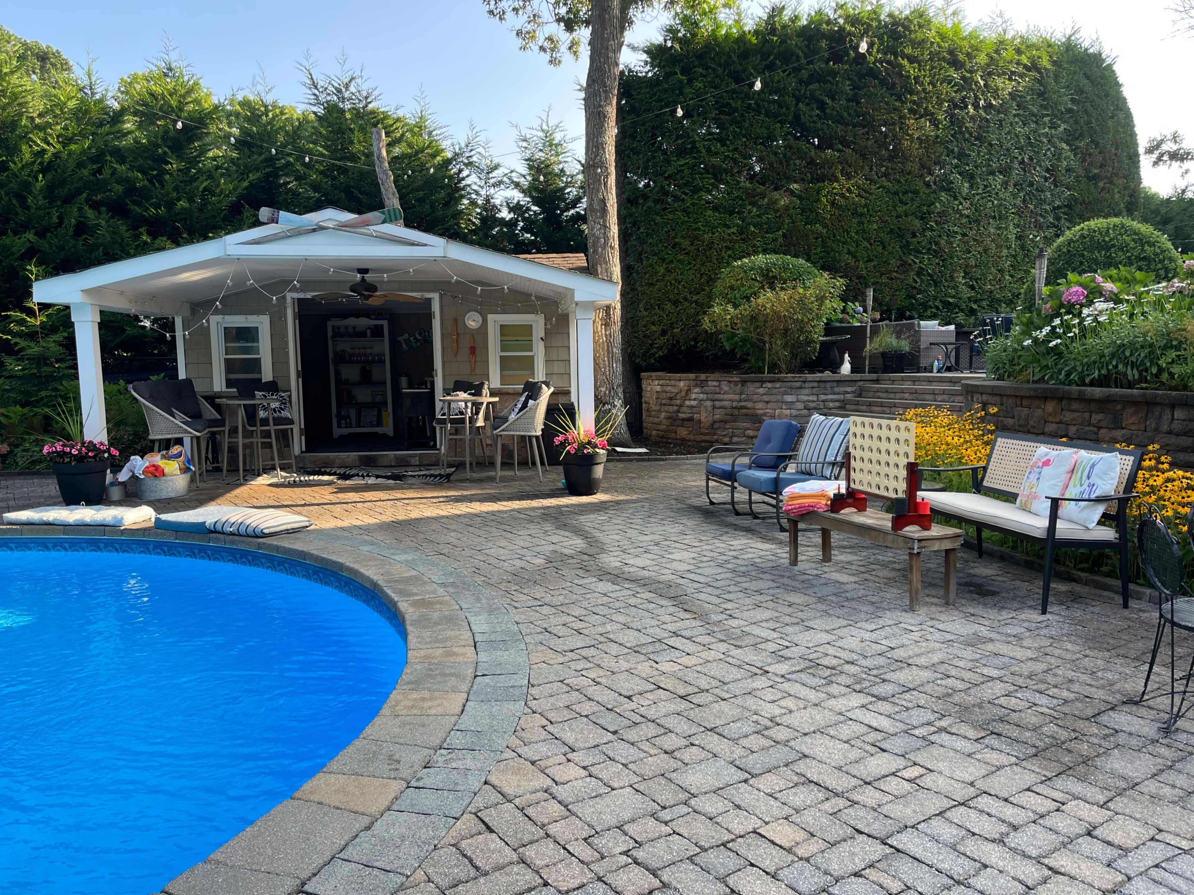 The image shows a landscaped backyard with a pool, a small outdoor shed, and various seating areas surrounded by greenery and flower beds.