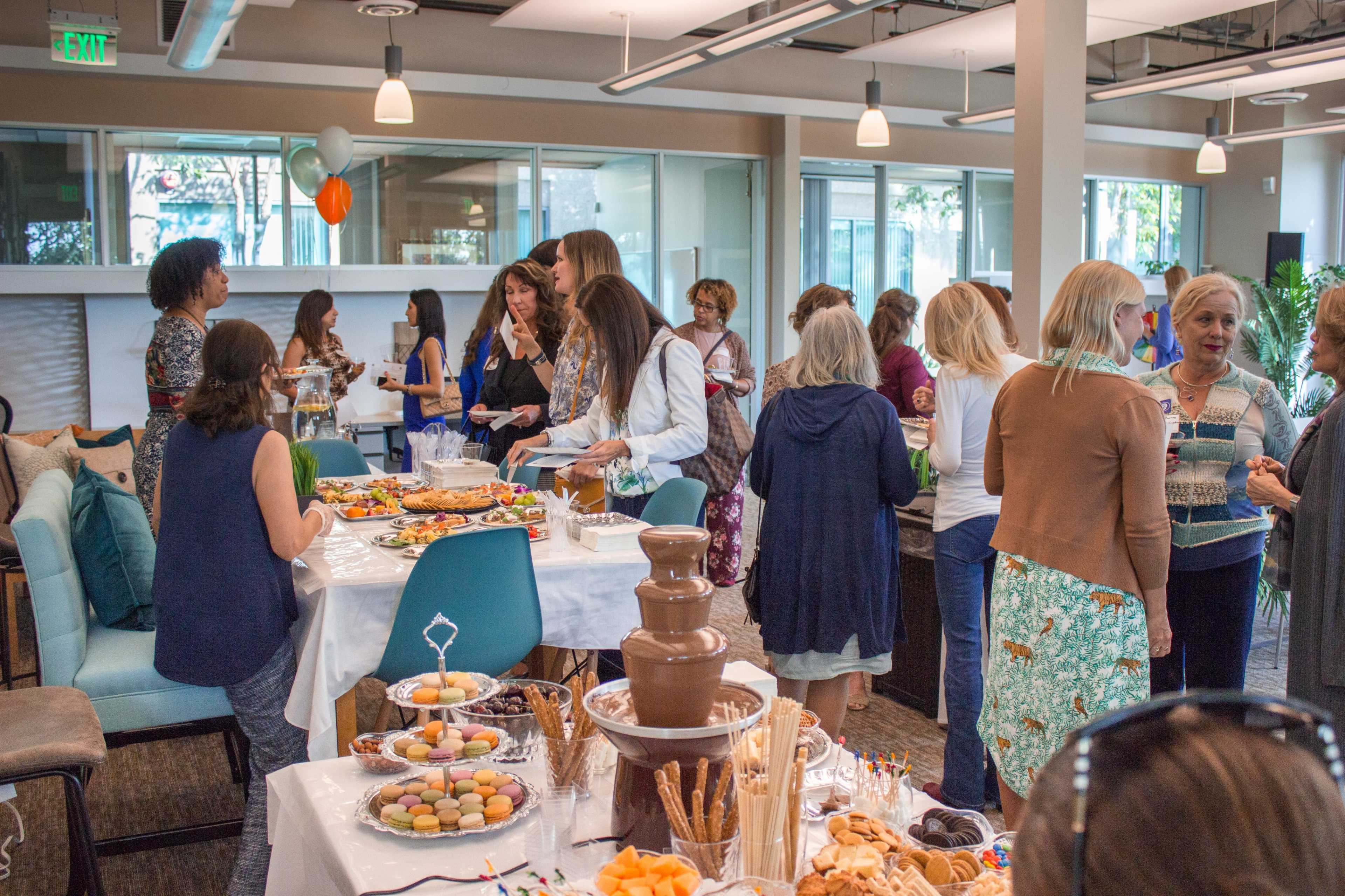 A group of people mingle at a social gathering featuring a buffet table with various foods and a chocolate fountain.