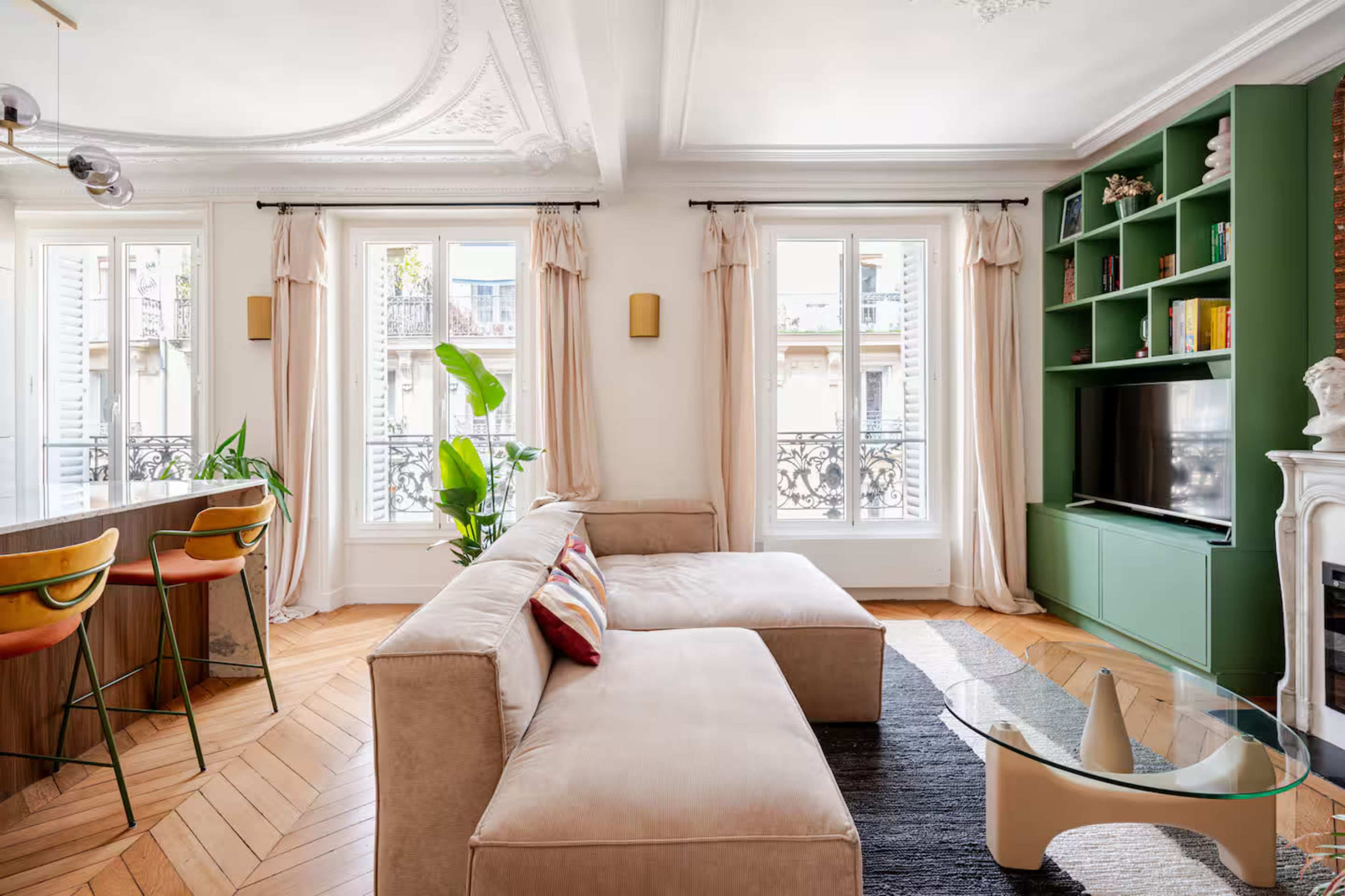 The image shows a well-lit living room featuring a beige sectional sofa, a green bookshelf, and large windows with beige curtains.