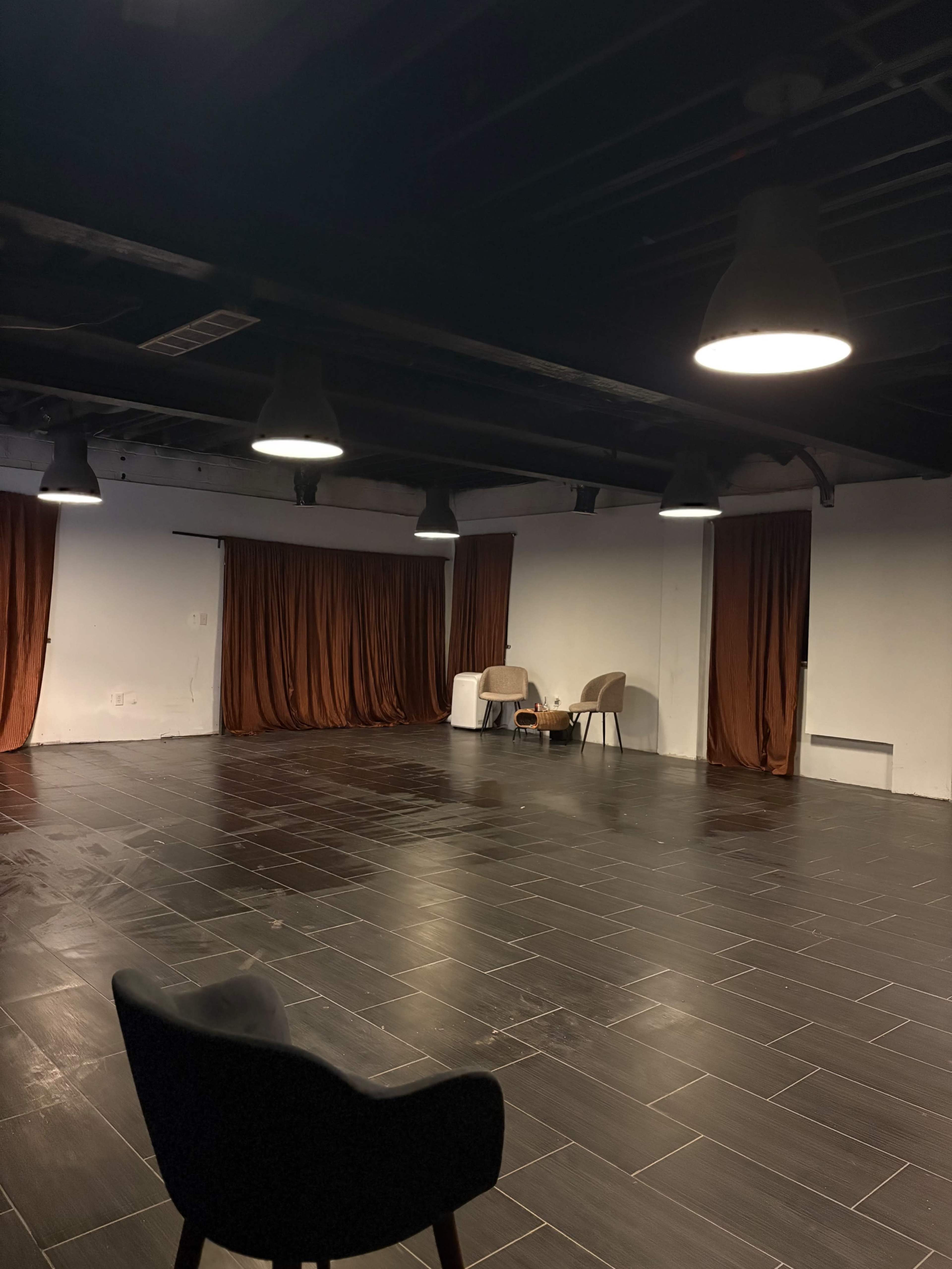 The image shows an empty room with dark tiled flooring, two brown curtains, and a small table with two chairs under overhead lights.