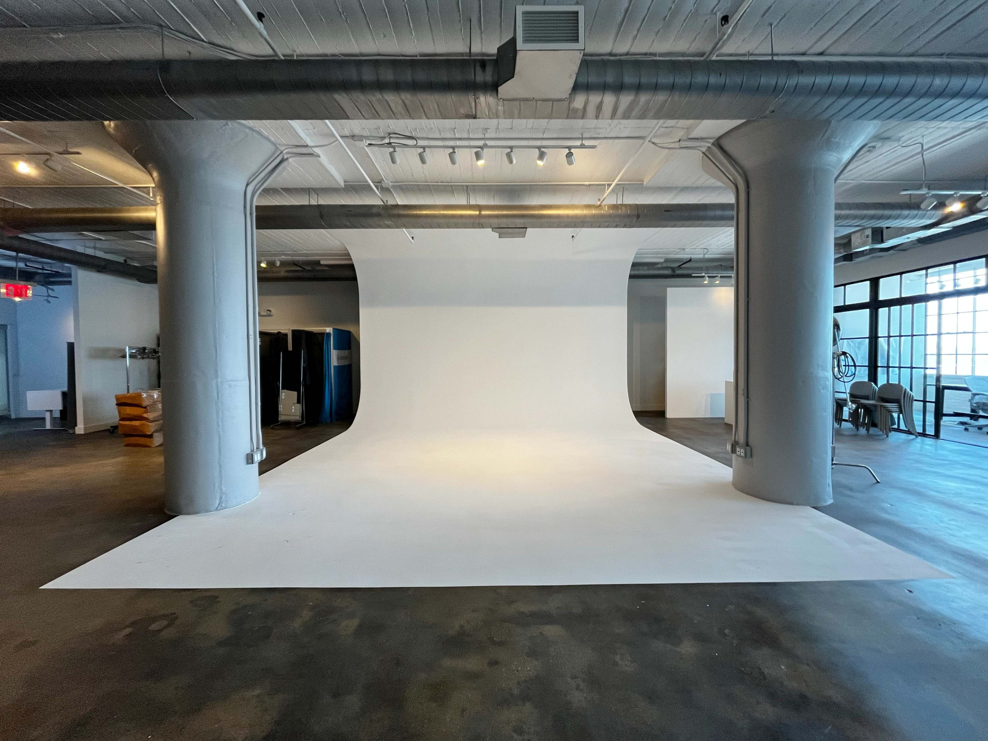 A spacious studio with a white backdrop and concrete floors, featuring large pillars and minimal furniture.