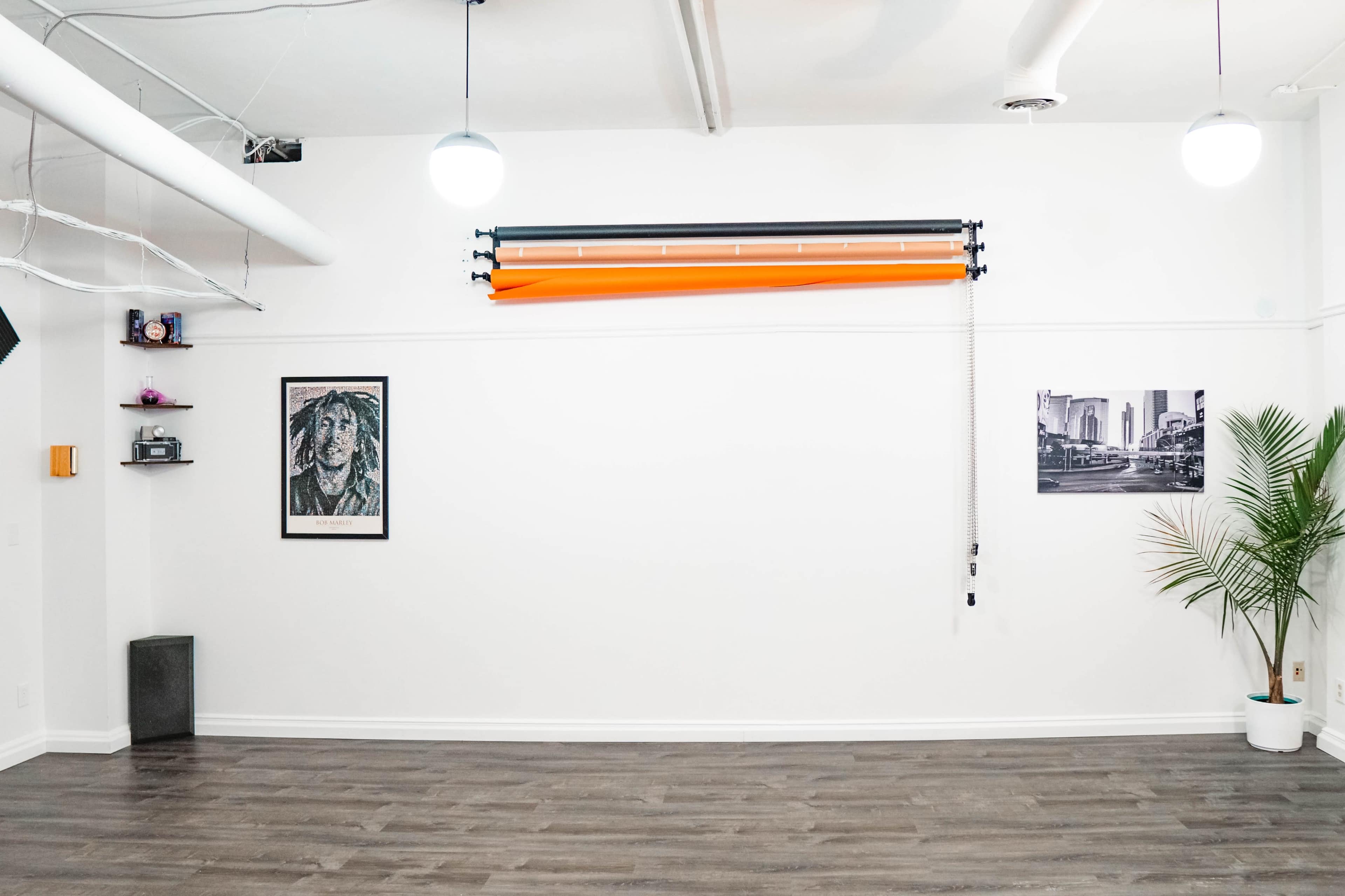 A spacious, empty room with wooden flooring, featuring a black and white photo on one wall, a framed portrait on another, and an orange roll-up backdrop mounted above.