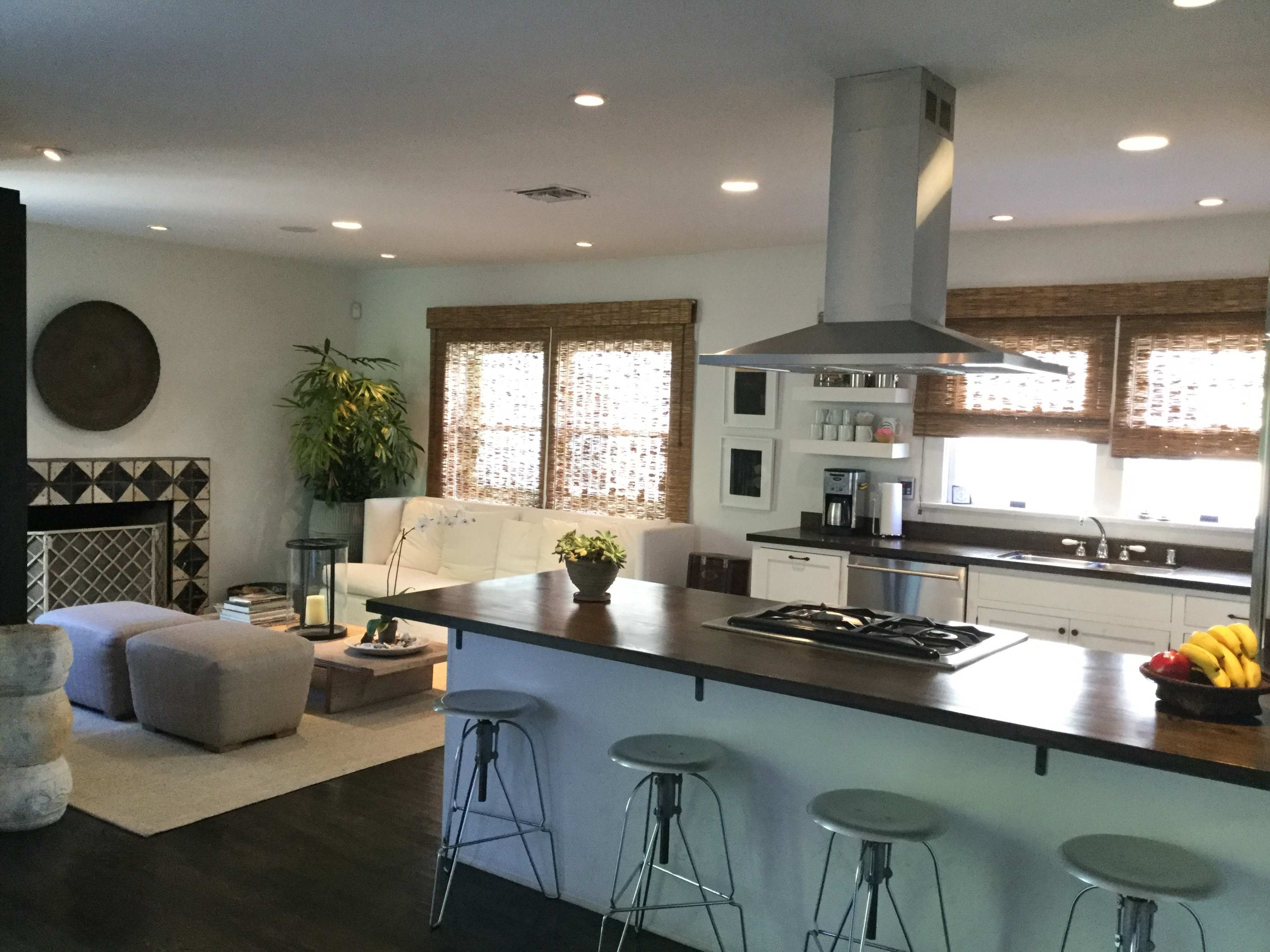 A modern kitchen and living area features a central island with bar stools, a comfortable seating arrangement, and large windows adorned with woven shades.