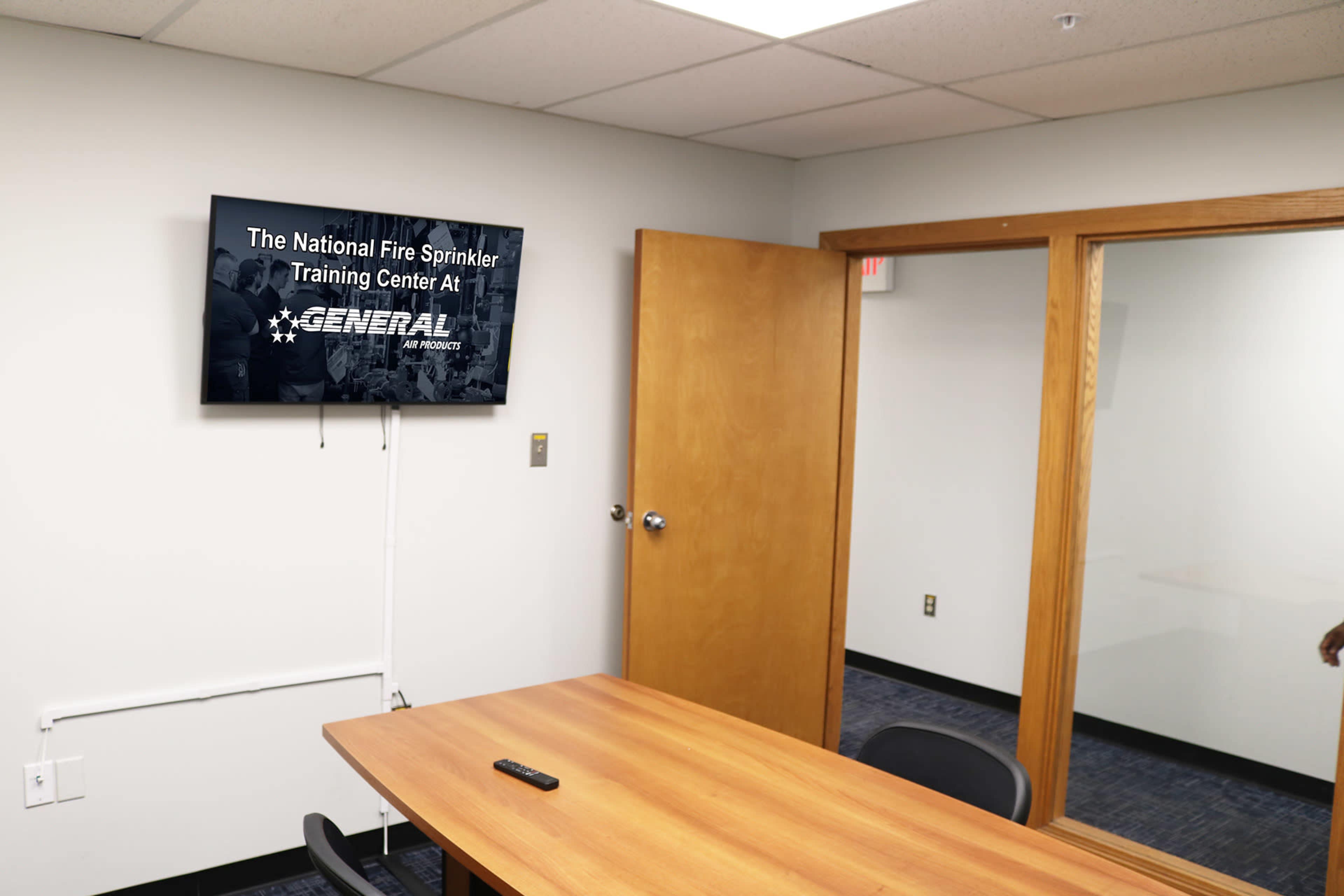 A conference room features a wooden table, a television mounted on the wall displaying "The National Fire Sprinkler Training Center Art," and a glass wall.