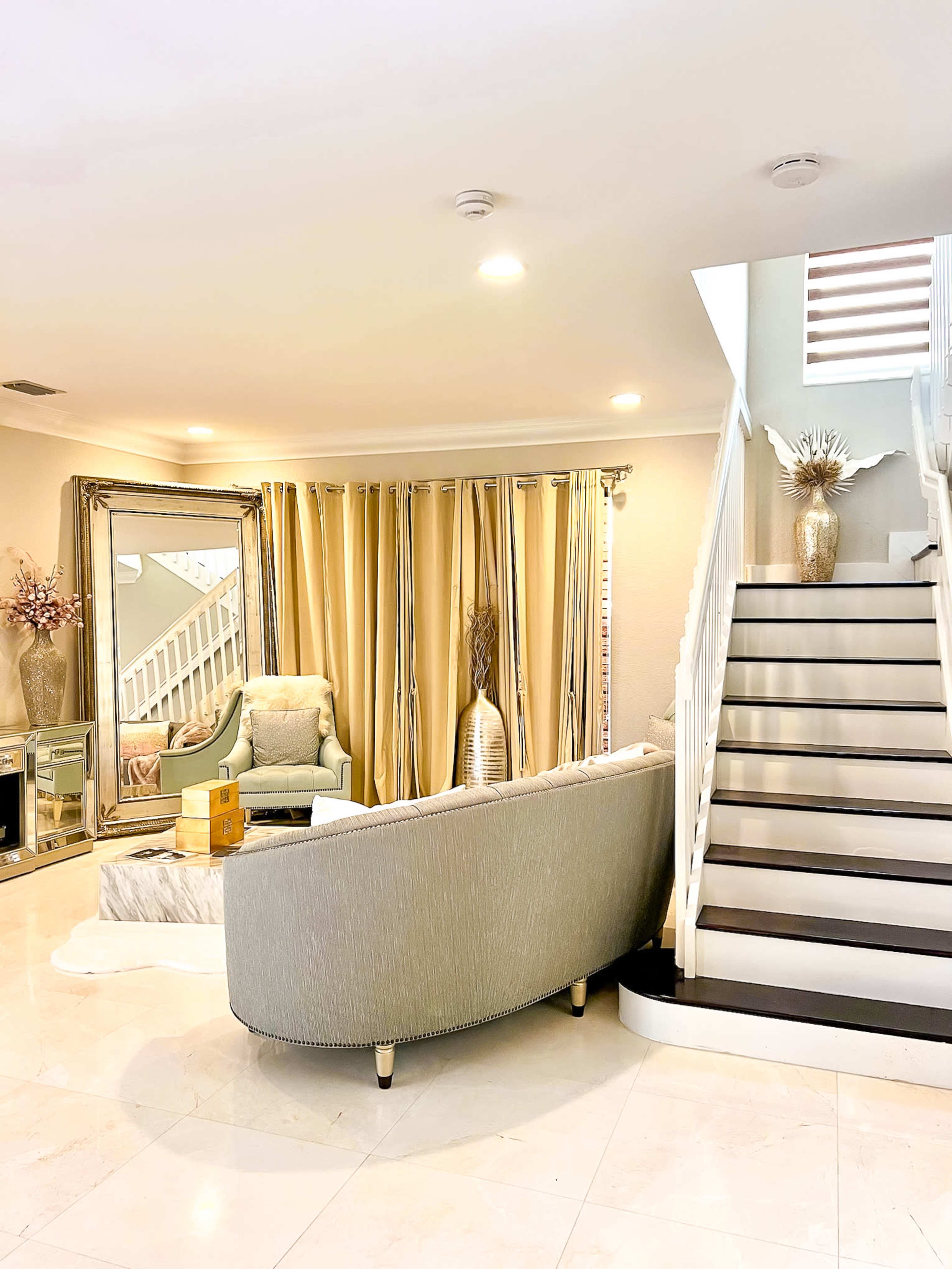 The image shows a modern living room with a curved sofa, decorative curtains, and a staircase leading to the upper floor.