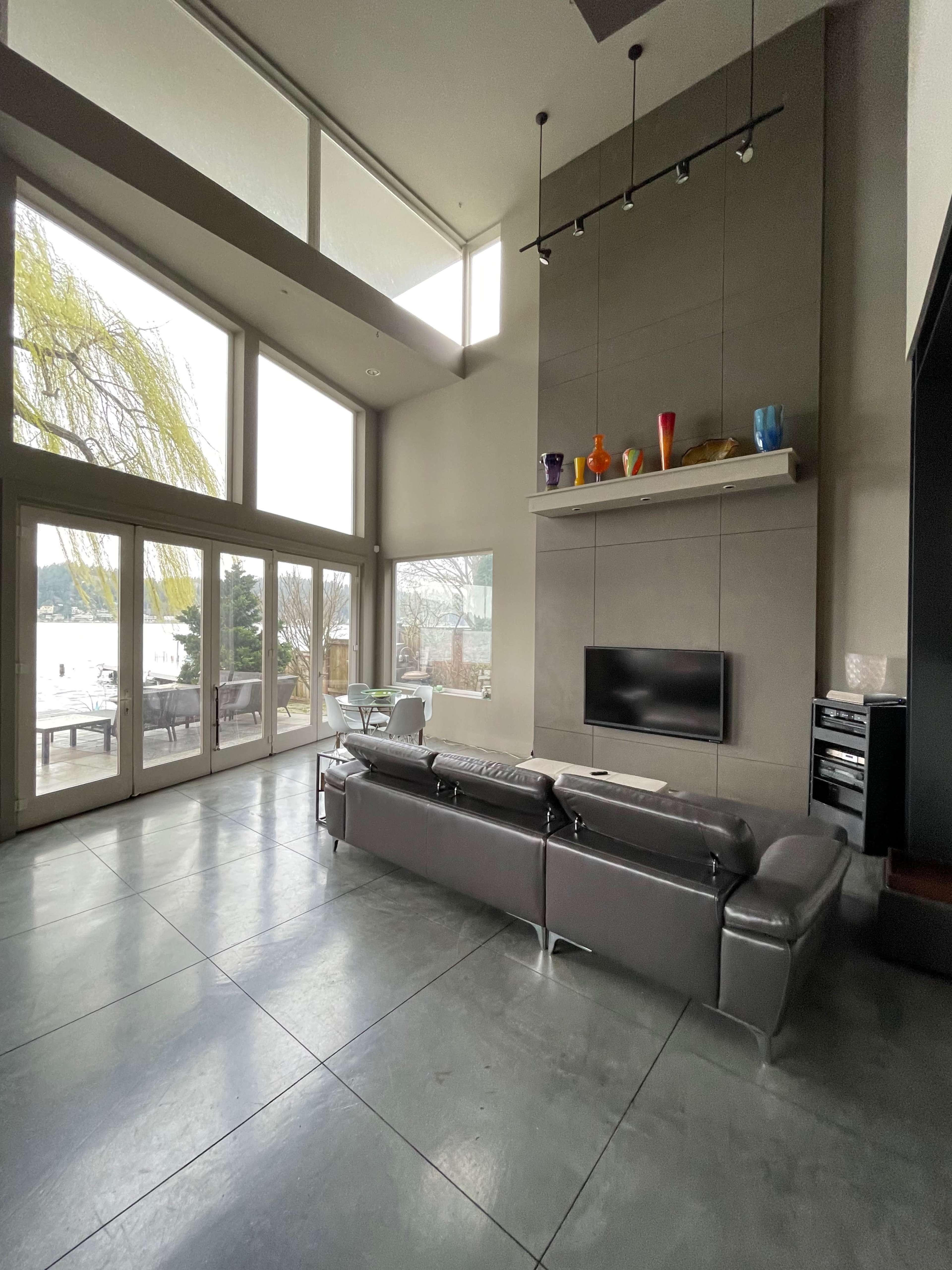 The image shows a modern living room with large windows overlooking a lake, featuring a gray sectional sofa and a mounted television on a stone wall.
