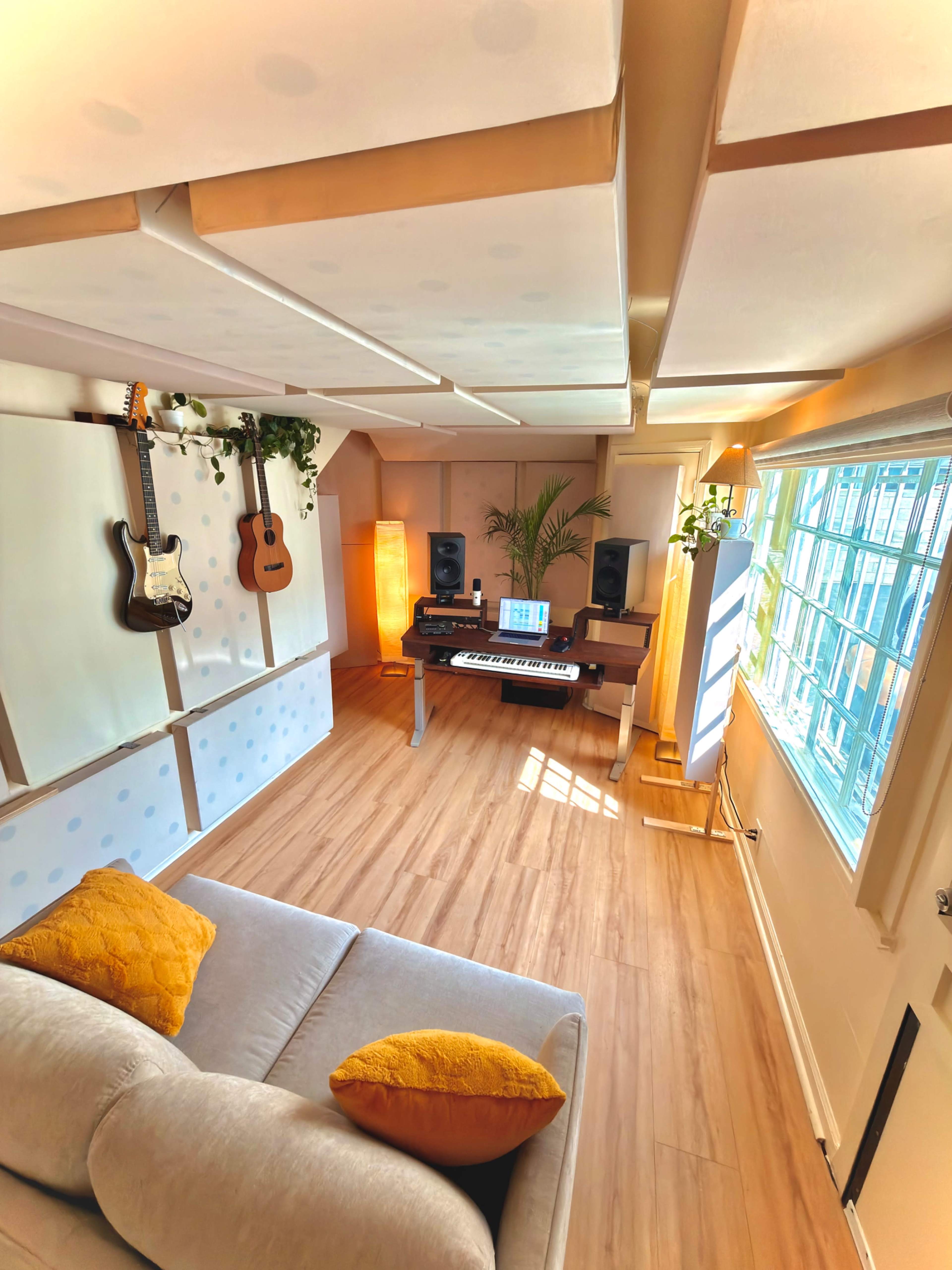 The image shows a cozy music studio featuring a keyboard, speakers, guitars on the wall, and a sofa with yellow pillows.