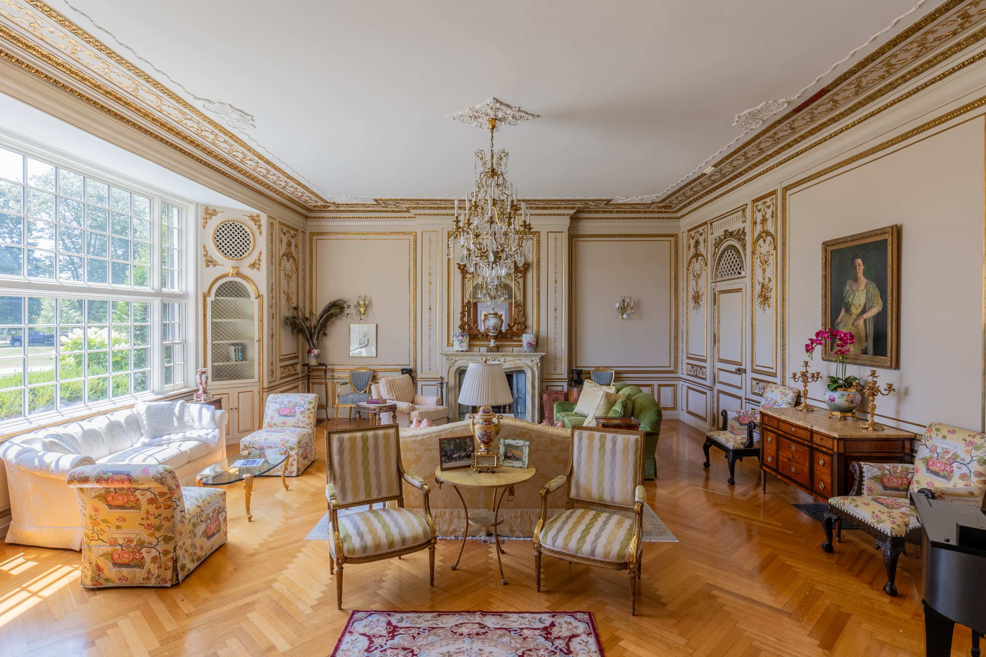 The room features ornate decor with a chandelier, floral-patterned furniture, and a fireplace, complemented by large windows letting in natural light.