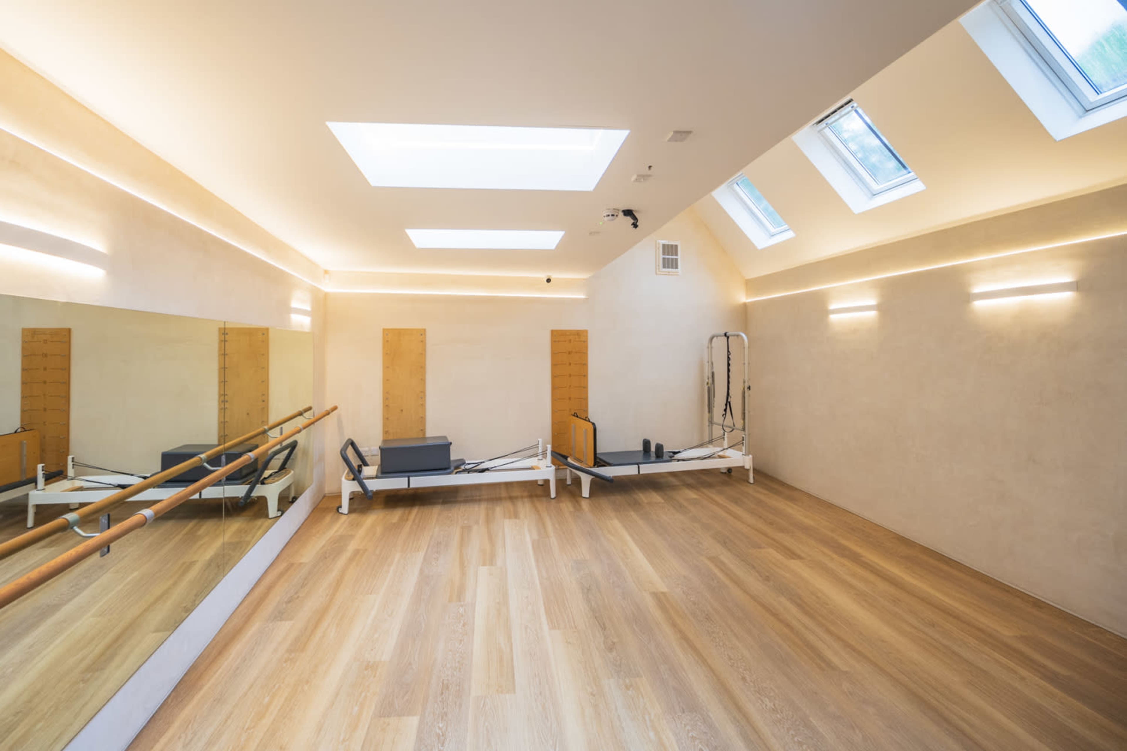 The image shows a bright, modern studio with wooden floors, large skylights, and a reformer Pilates machine near a wall mirror.