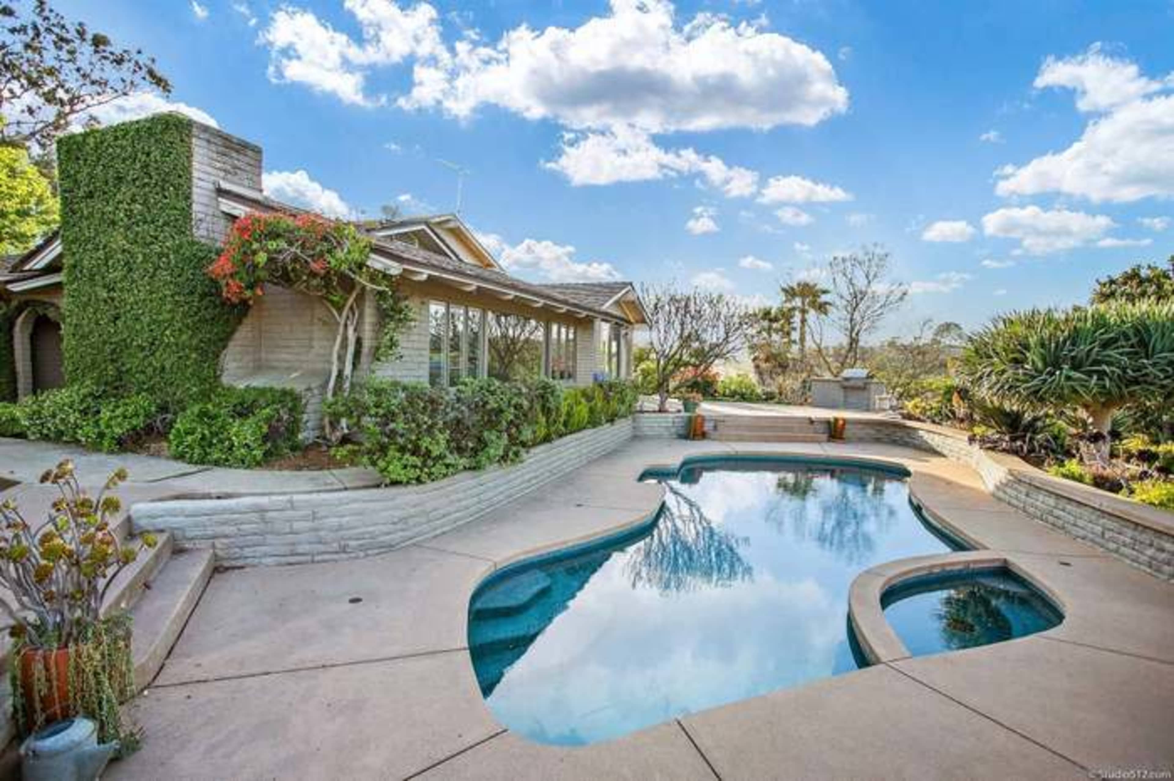 A swimming pool with a curved shape is surrounded by landscaped gardens and a house with large windows and a stone facade under a partly cloudy sky.