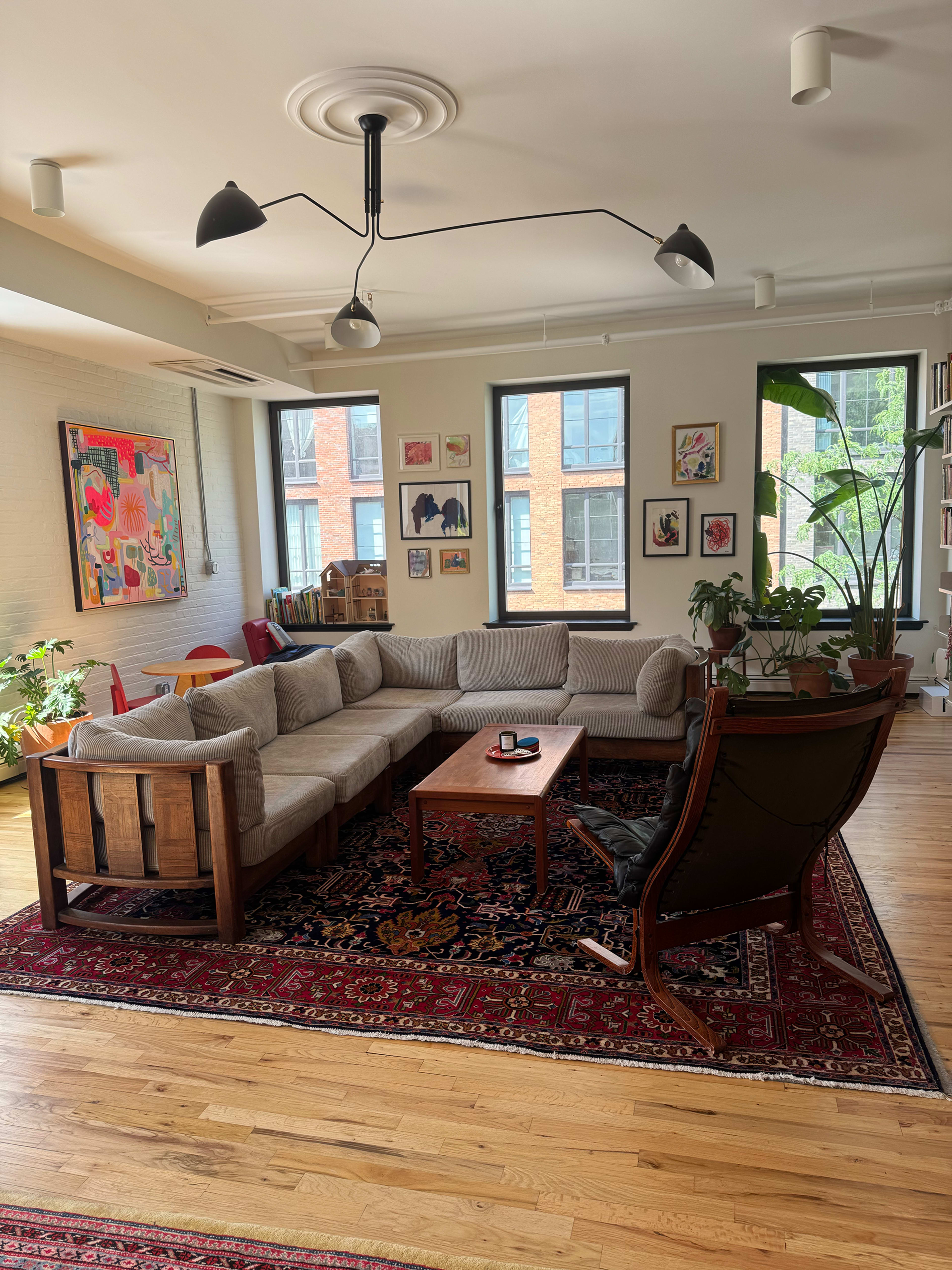 The image shows a spacious living room with a large sectional couch, a wooden coffee table in the center, and large windows letting in natural light.