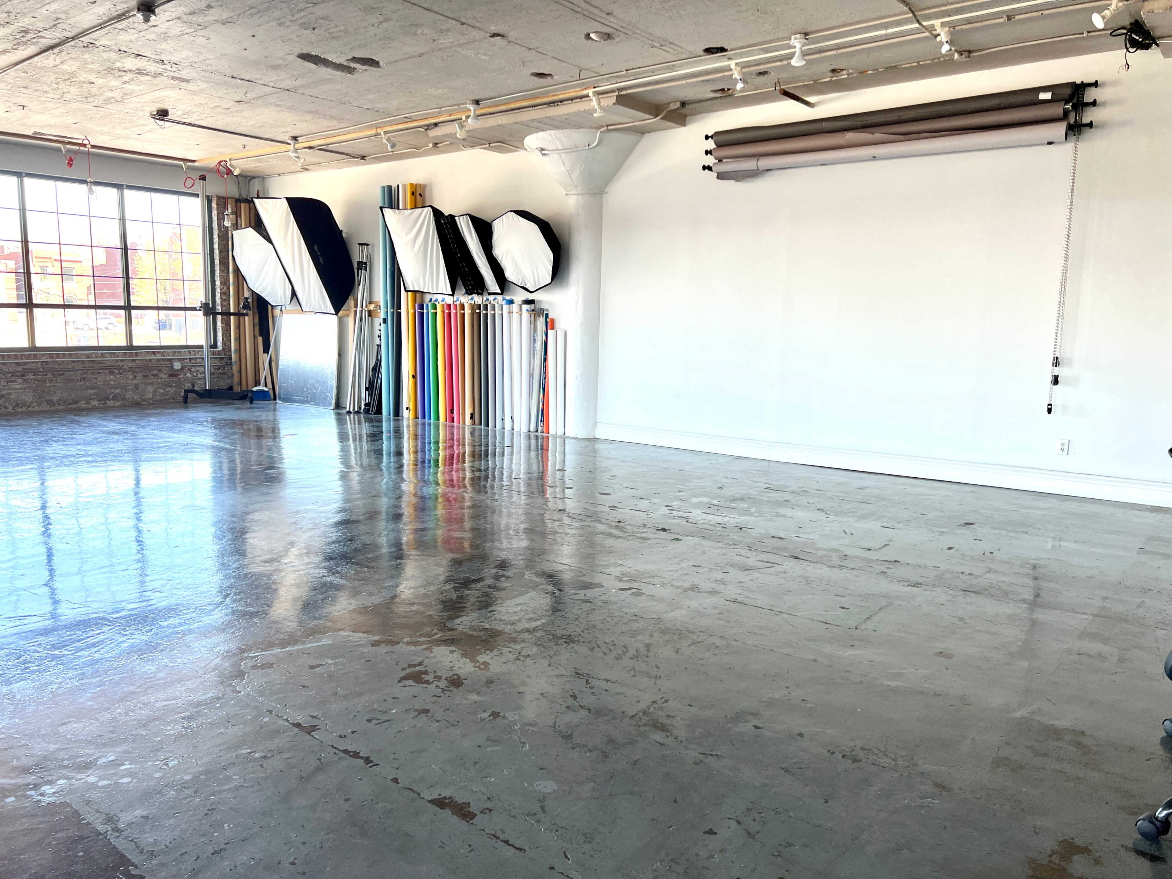 The image shows an empty studio space featuring a polished concrete floor, large windows, and a backdrop setup with various fabric colors neatly arranged against a white wall.