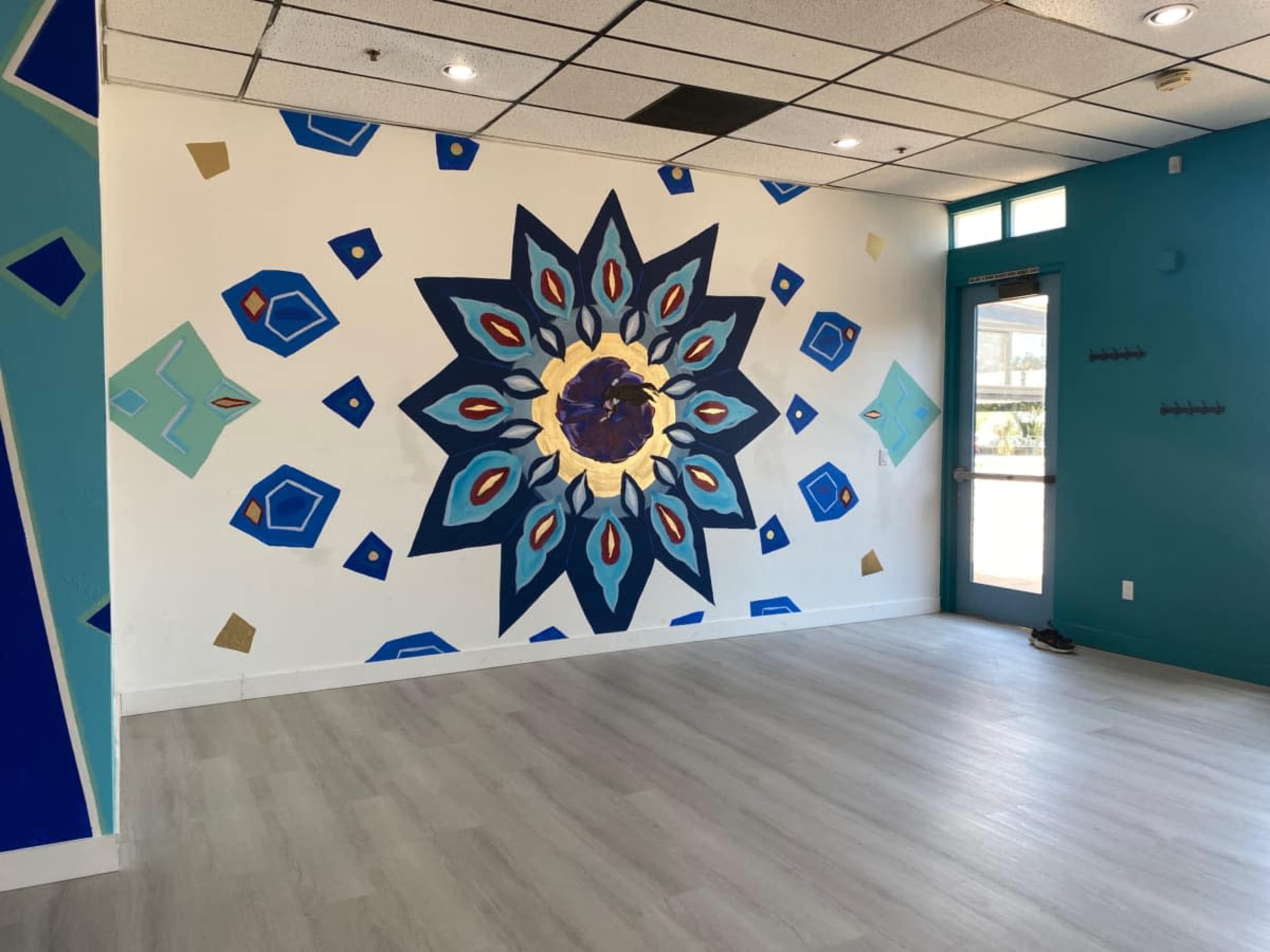 The image shows an interior space with a large, colorful geometric mural on one wall, featuring a central design with shades of blue, red, and gold, and light-colored flooring.