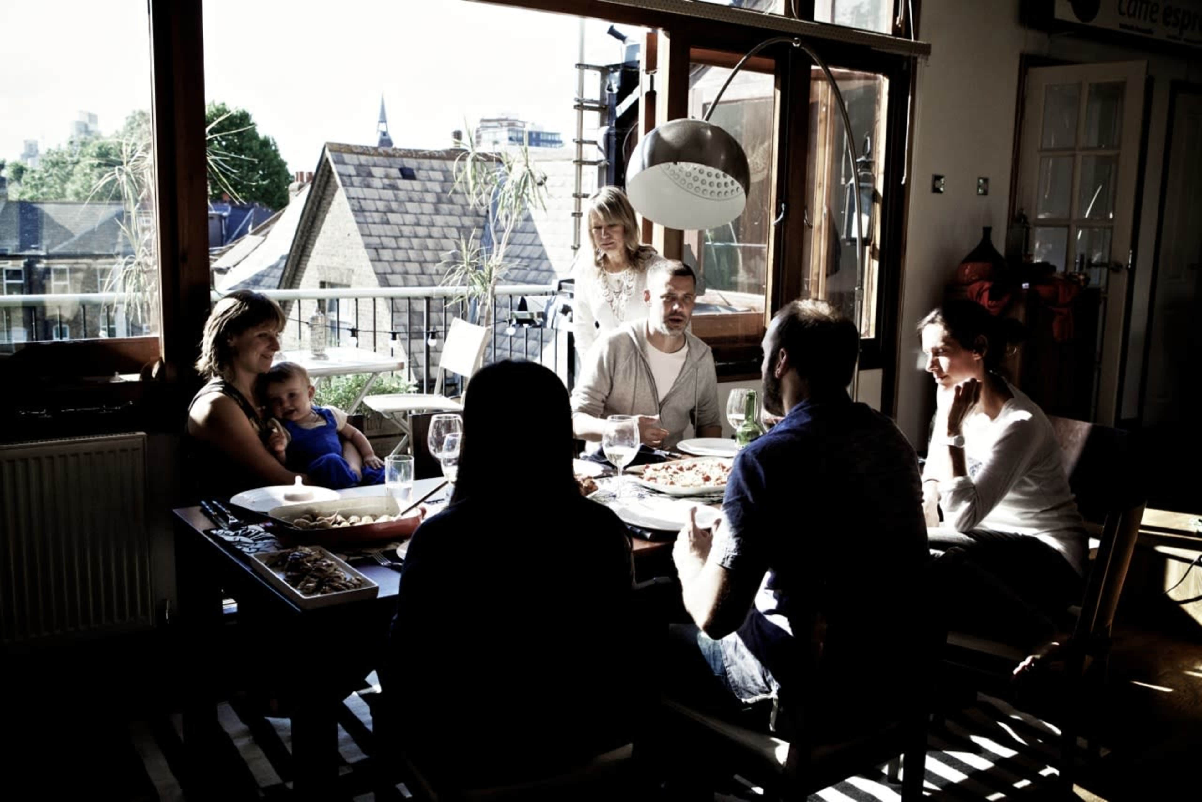 A group of six people sits around a table in a sunlit room, sharing food and conversation.