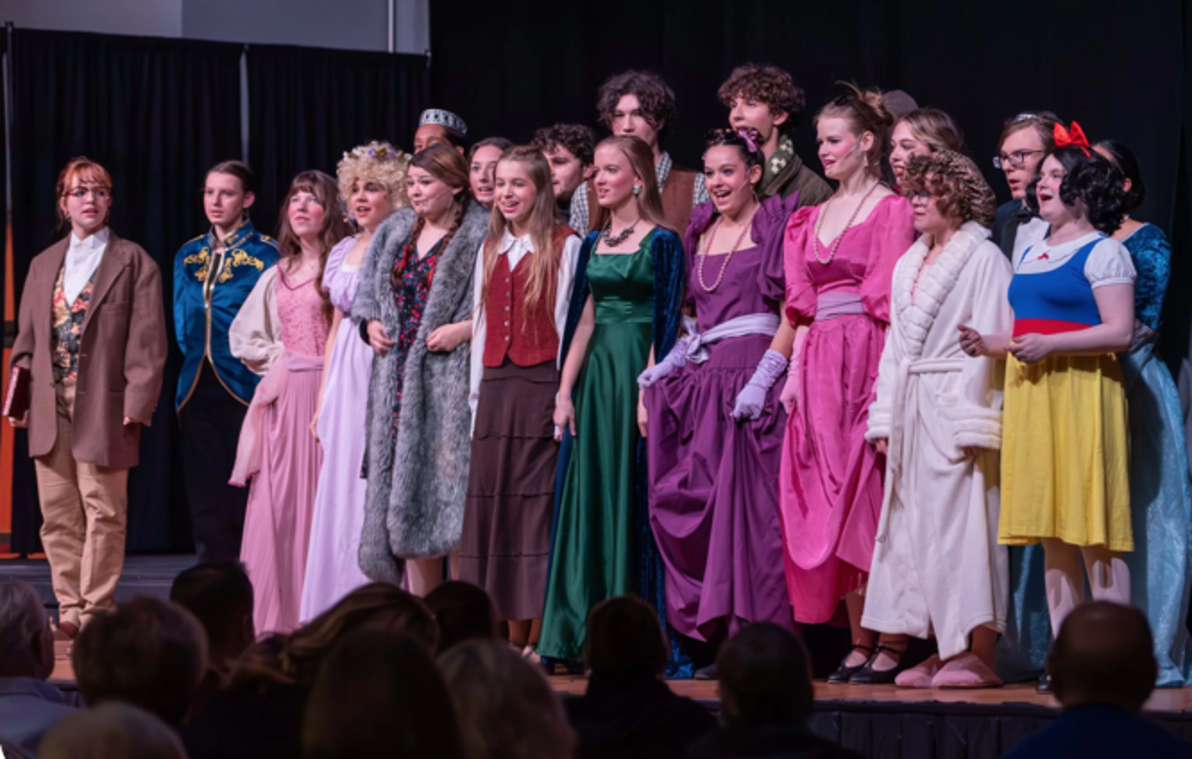 A group of actors dressed in various colorful costumes stands on stage, posing together during a performance.