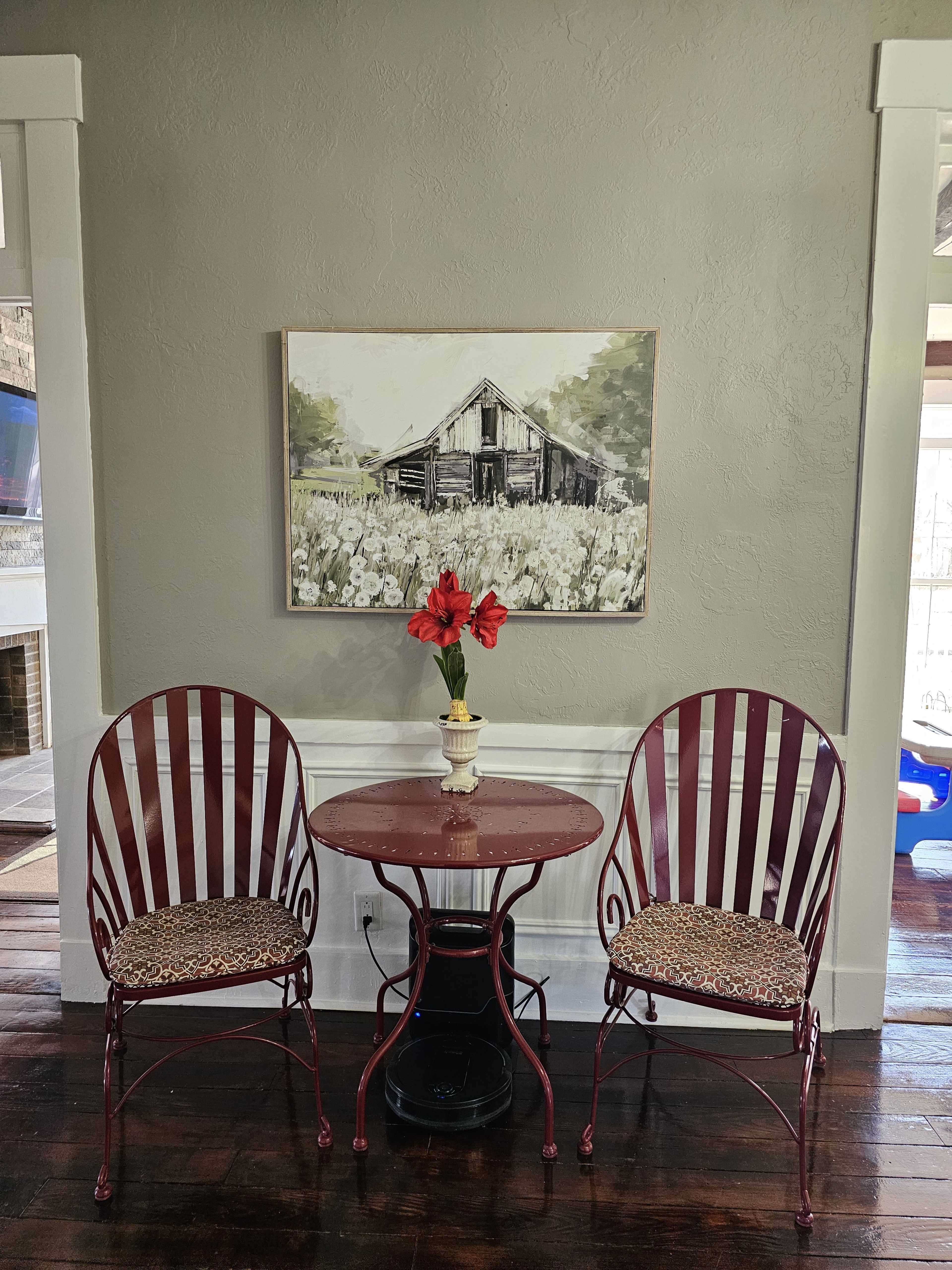 A small round table with two chairs is positioned against a gray wall, featuring a floral centerpiece and a painting of a barn above it.