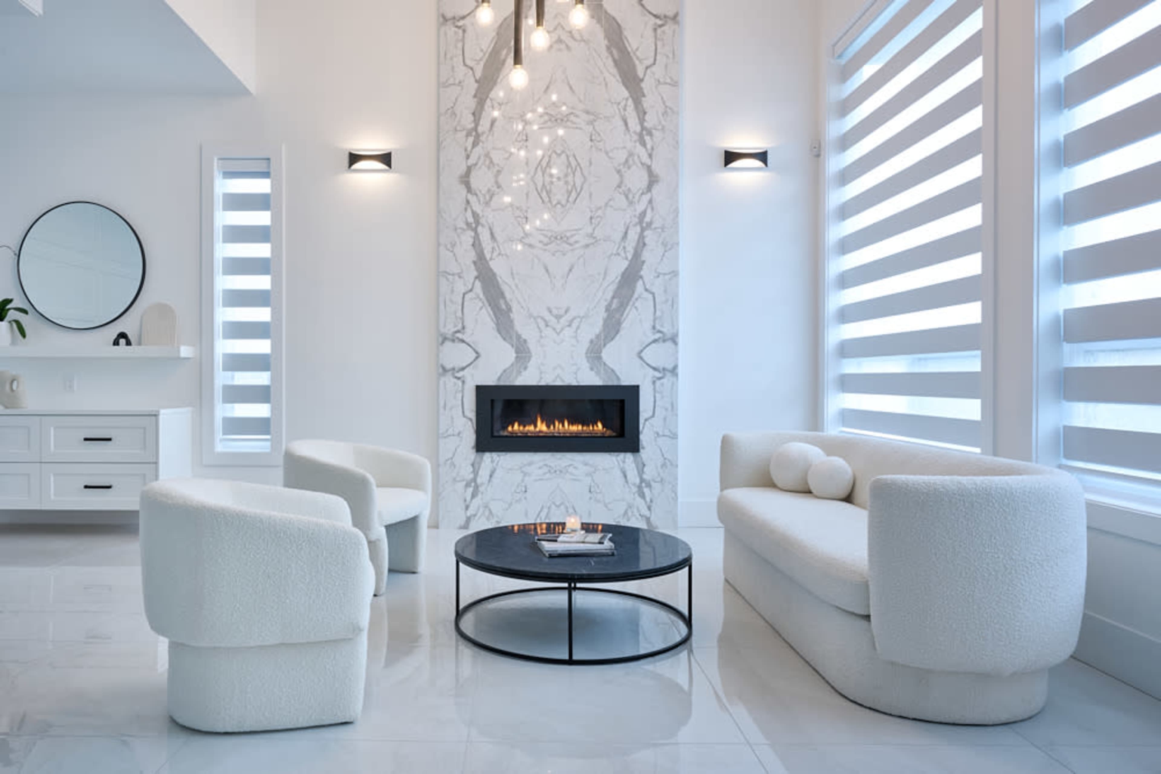 A modern living room features a white marble fireplace, a round black coffee table, and plush, white upholstered furniture.