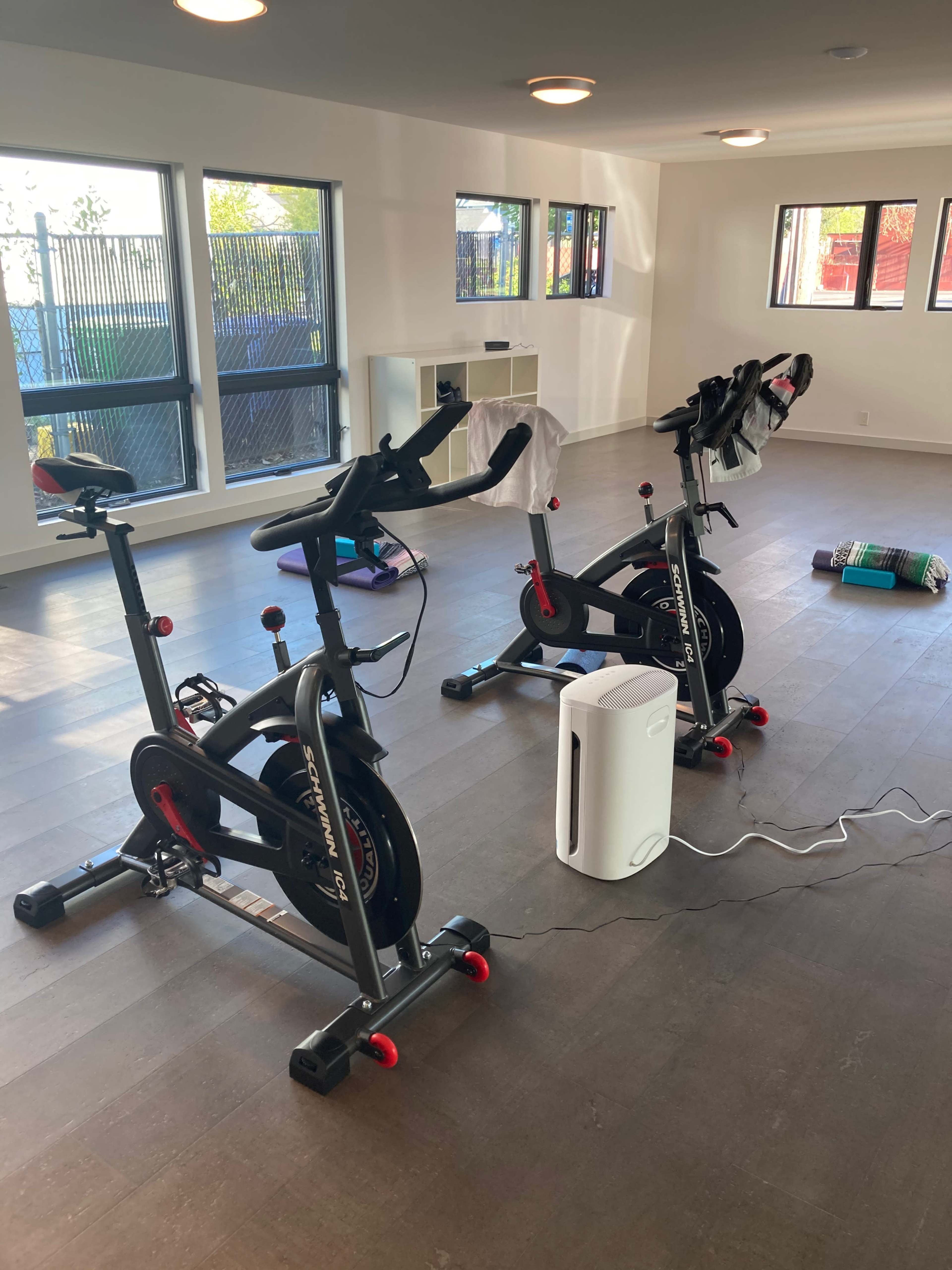 The image shows a fitness room with two stationary bicycles, a portable air purifier, and some workout mats on the floor.