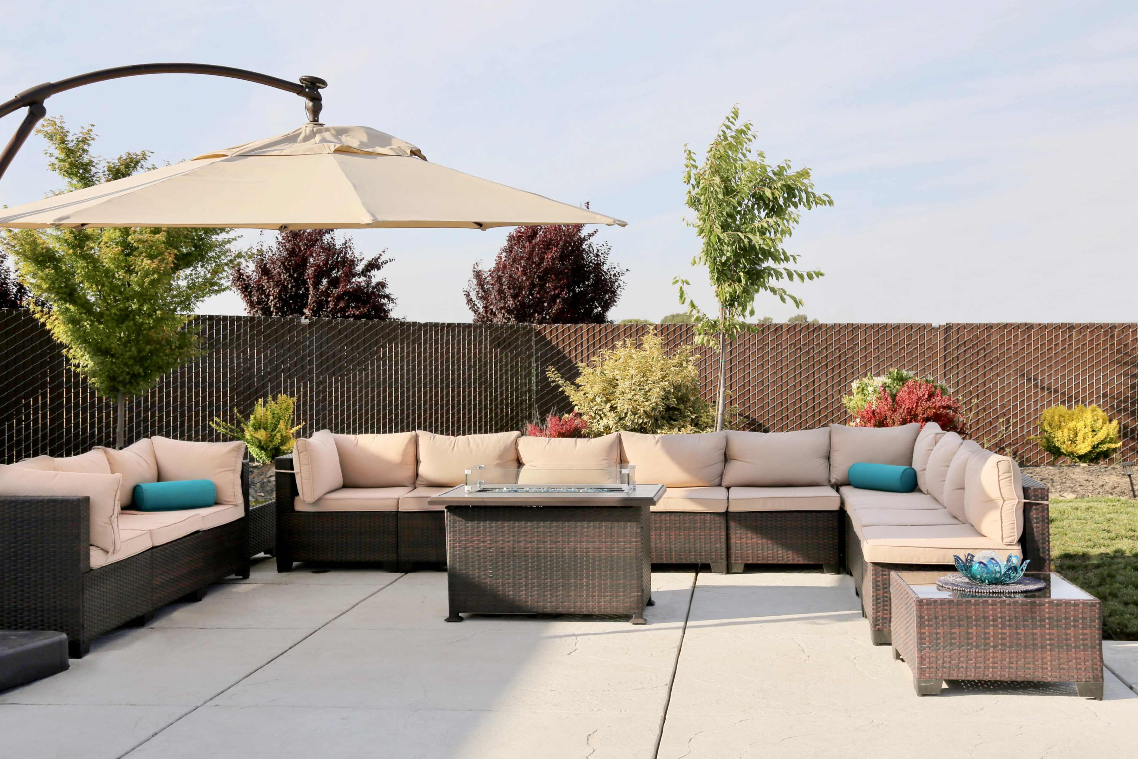 A patio area features a large sectional seating arrangement with neutral cushions, a glass-top coffee table, and an umbrella, surrounded by landscaped greenery.
