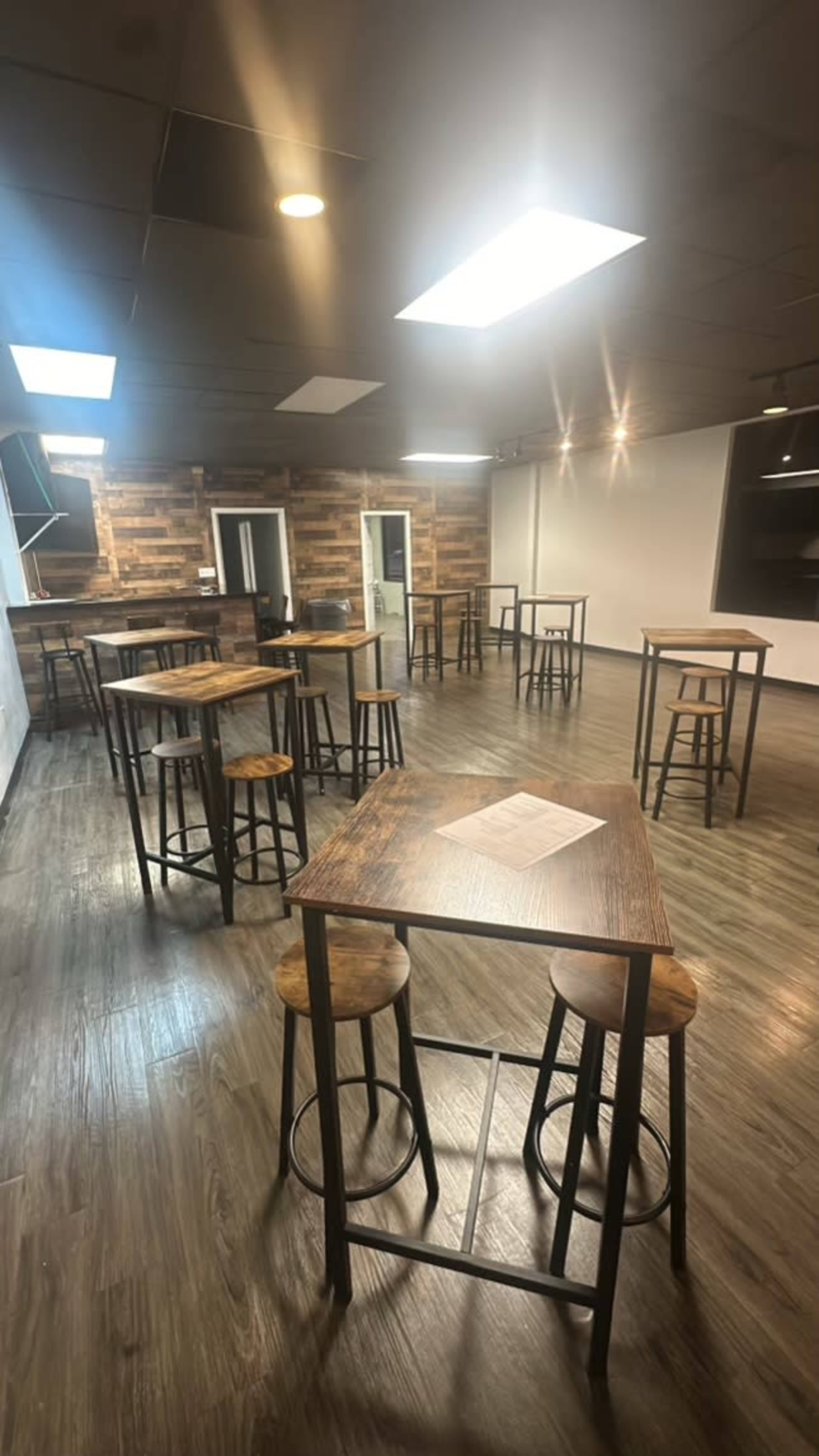 The room features several tall tables and stools arranged on a wooden floor, with large windows providing natural light from above.