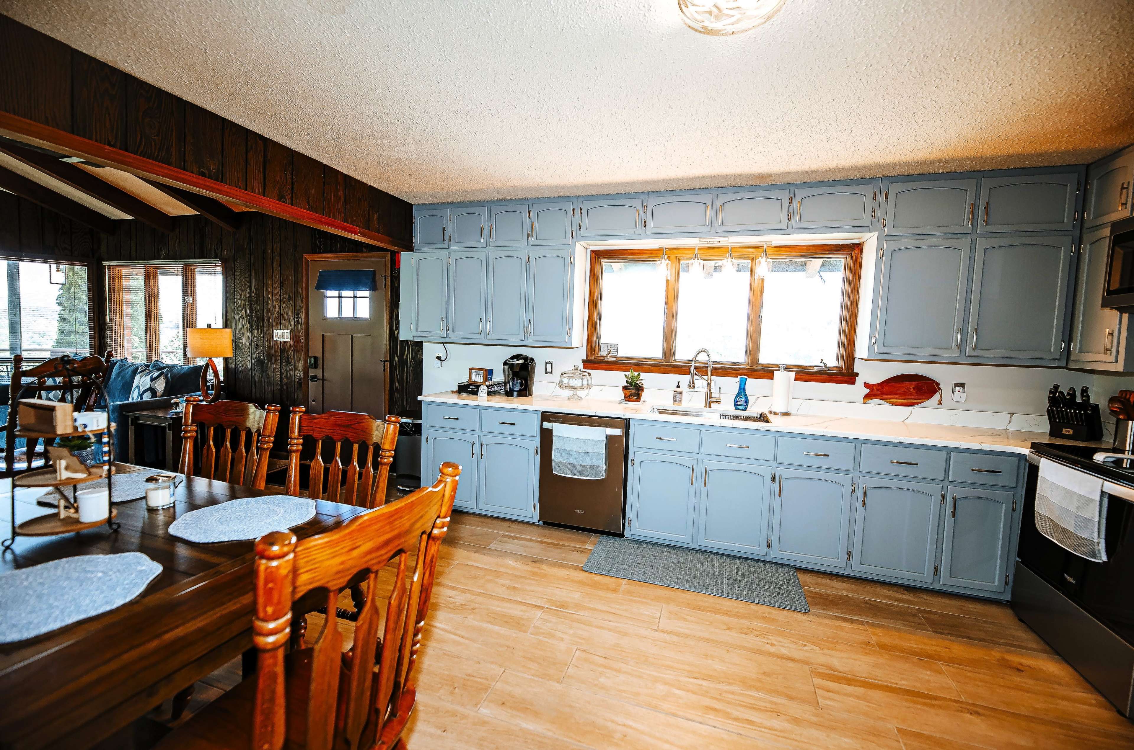 The image shows a residential kitchen with gray cabinets, a large window, and a wooden dining table with chairs.