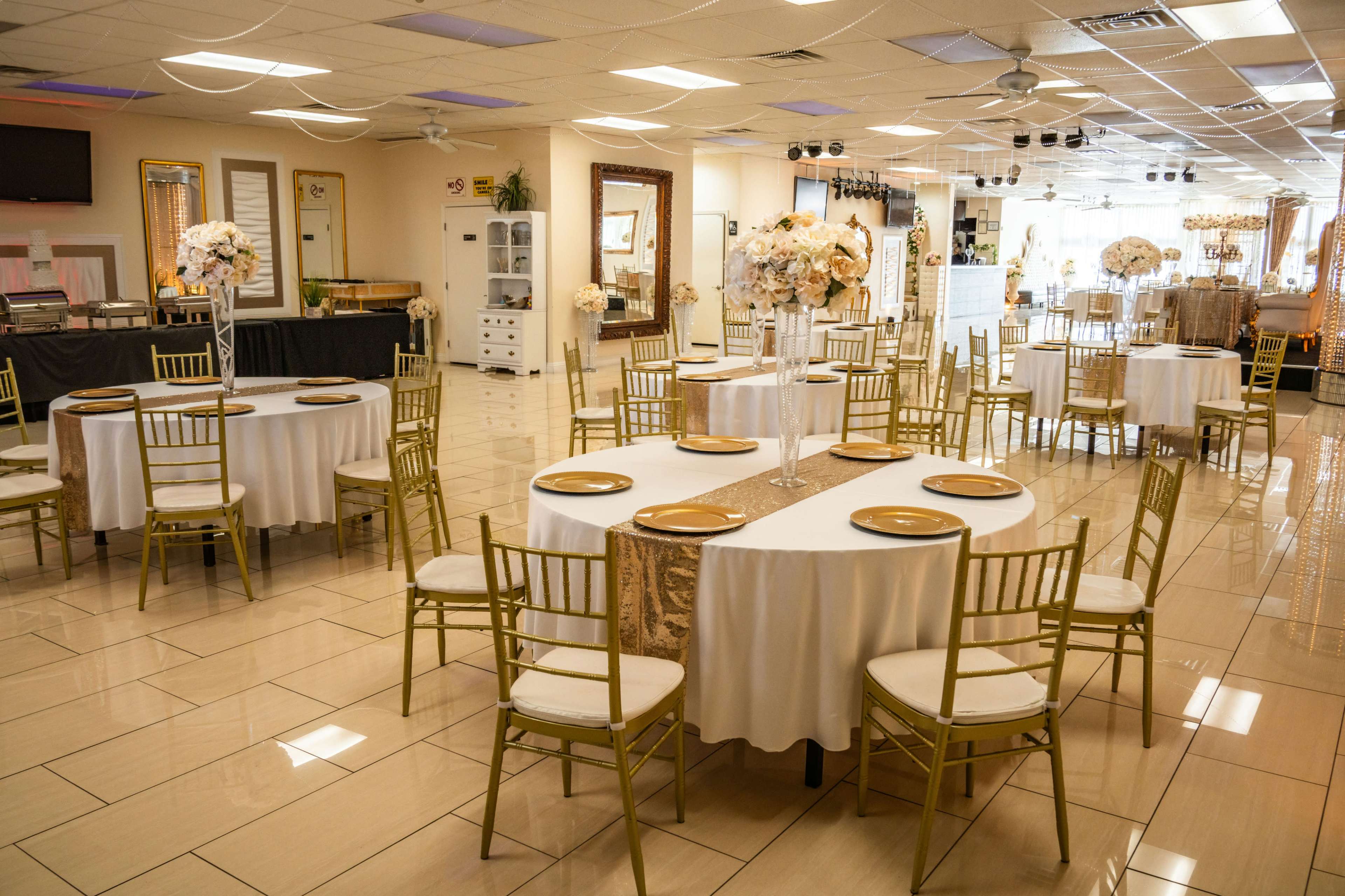 The image shows a spacious banquet hall arranged with round tables covered in white tablecloths, each set with gold plates and decorative centerpieces.